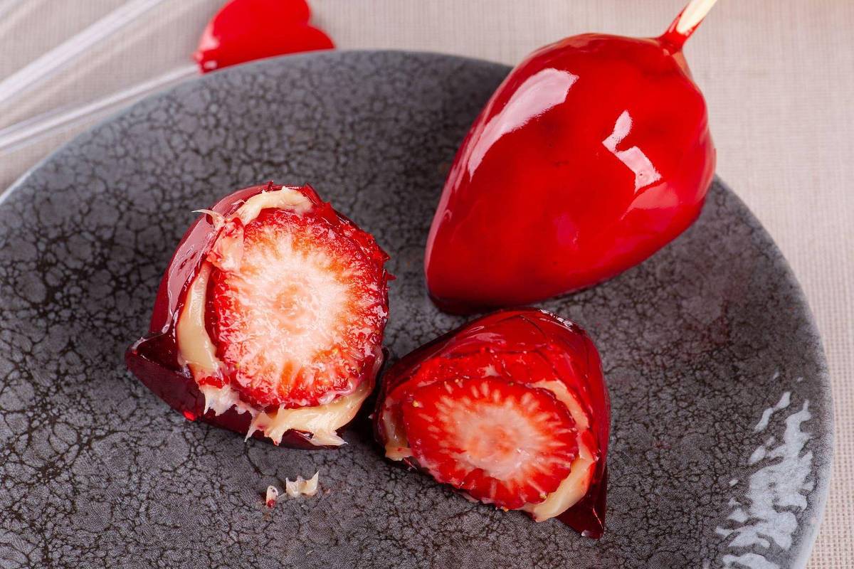 Um prato com um morango coberto com uma camada brilhante de chocolate vermelho, ao lado de dois morangos cortados ao meio, mostrando o interior cremoso e a polpa vermelha. O prato é de cor escura e a mesa tem uma toalha clara ao fundo.
