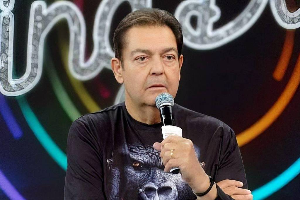 Um homem está em um estúdio de televisão, segurando um microfone. Ele tem cabelo castanho e está usando uma camiseta preta com uma estampa de um gorila. O fundo do estúdio é colorido, com luzes em tons de azul, verde, amarelo e vermelho, formando um círculo ao redor do homem. Ele parece sério e está com os braços cruzados.