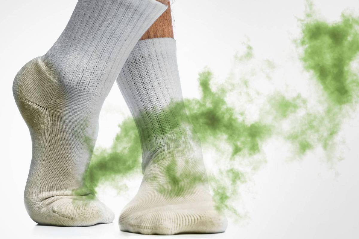 A imagem mostra um par de pés calçados com meias brancas. Há um efeito visual de fumaça verde saindo dos pés, sugerindo um odor desagradável. O fundo é claro, destacando os pés e o efeito de fumaça.