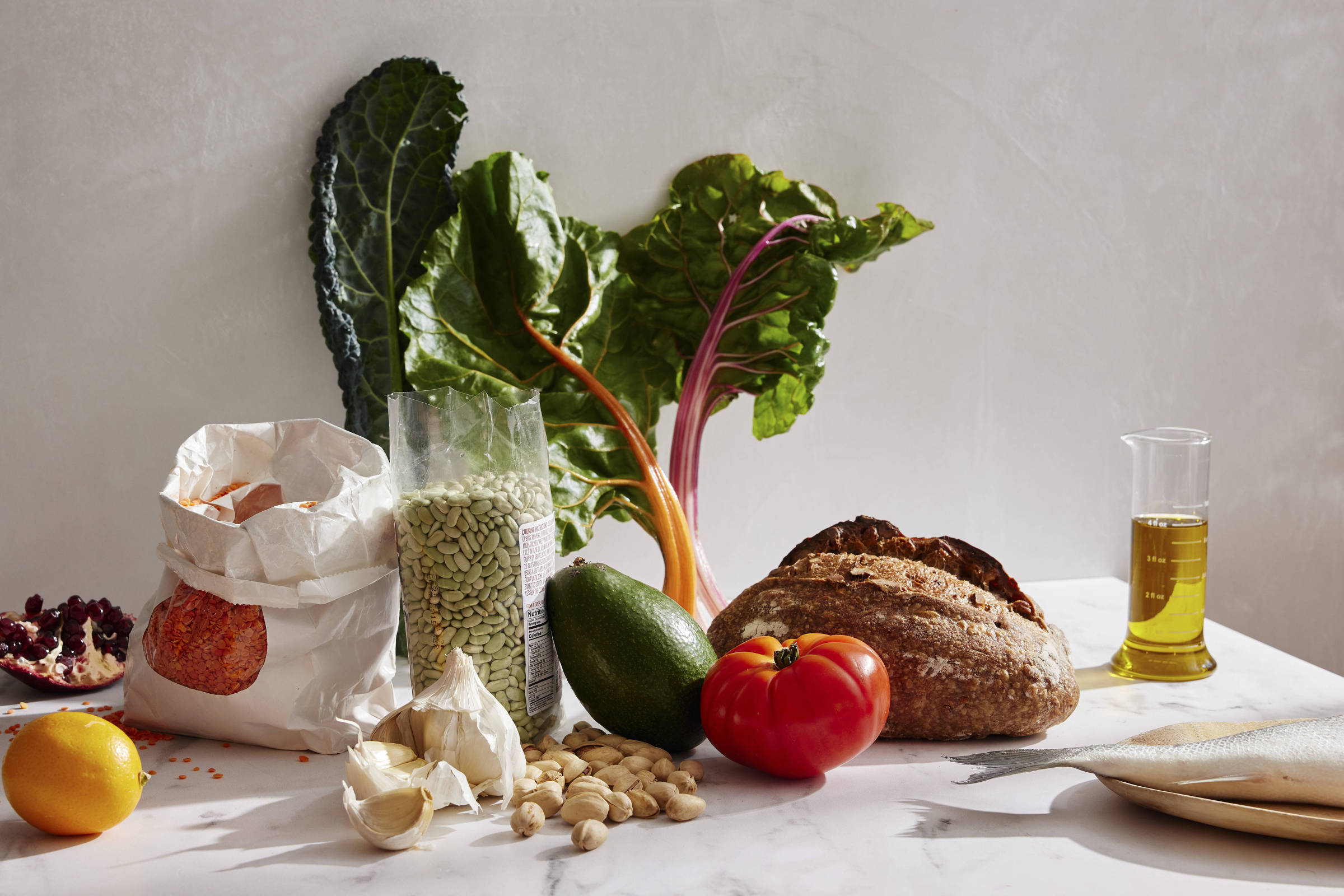 A imagem mostra uma variedade de ingredientes frescos dispostos sobre uma mesa. Há um saco de farinha, um recipiente com lentilhas verdes, um abacate, um tomate vermelho, um punhado de amêndoas, dentes de alho, uma fatia de pão integral, um limão, e folhas de acelga. Também há um frasco de azeite ao lado dos ingredientes.