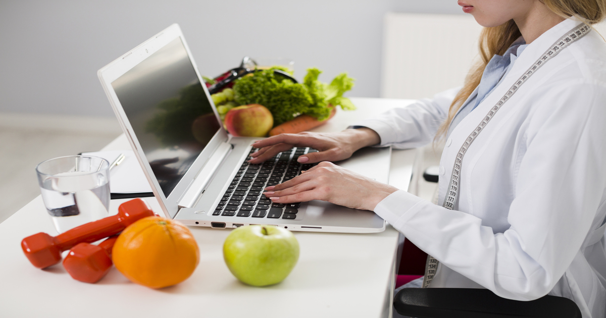 Imagem de uma mulher vestida de branco digitando num computador, enquanto ao lado deste, de ambos os lados, encontram-se alimentos como maçãs e verduras e também pequenos pesos usados em musculação