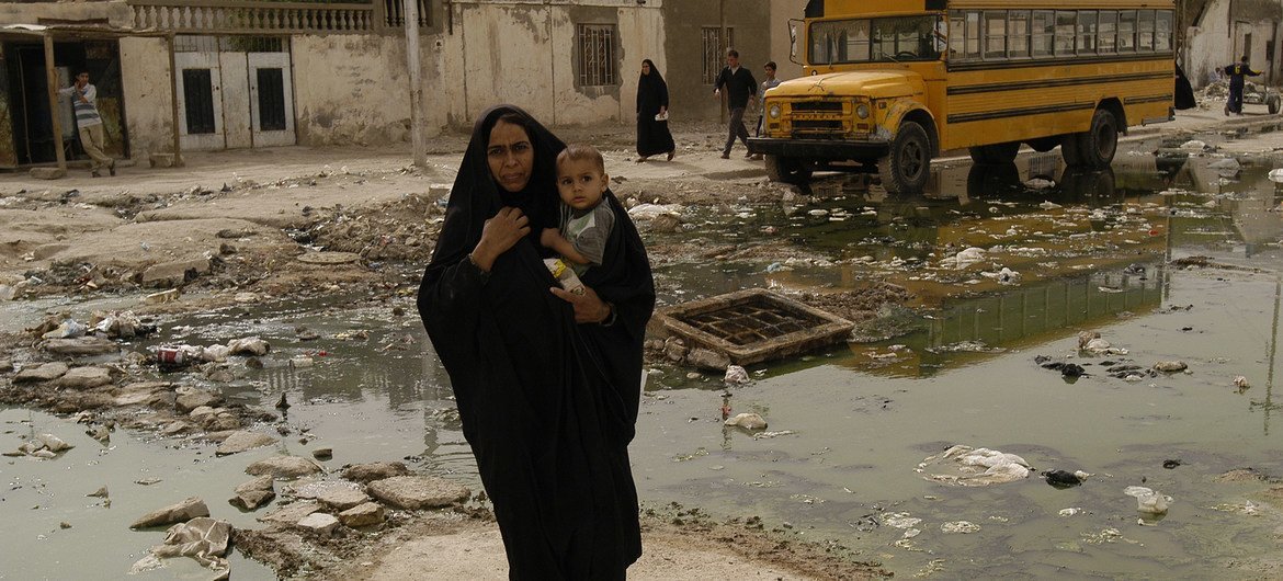 Uma mulher carregando uma criança atravessa uma rua coberta de lixo, a caminho de um centro de saúde primário, na cidade iraquiana de Basra (foto de arquivo)
