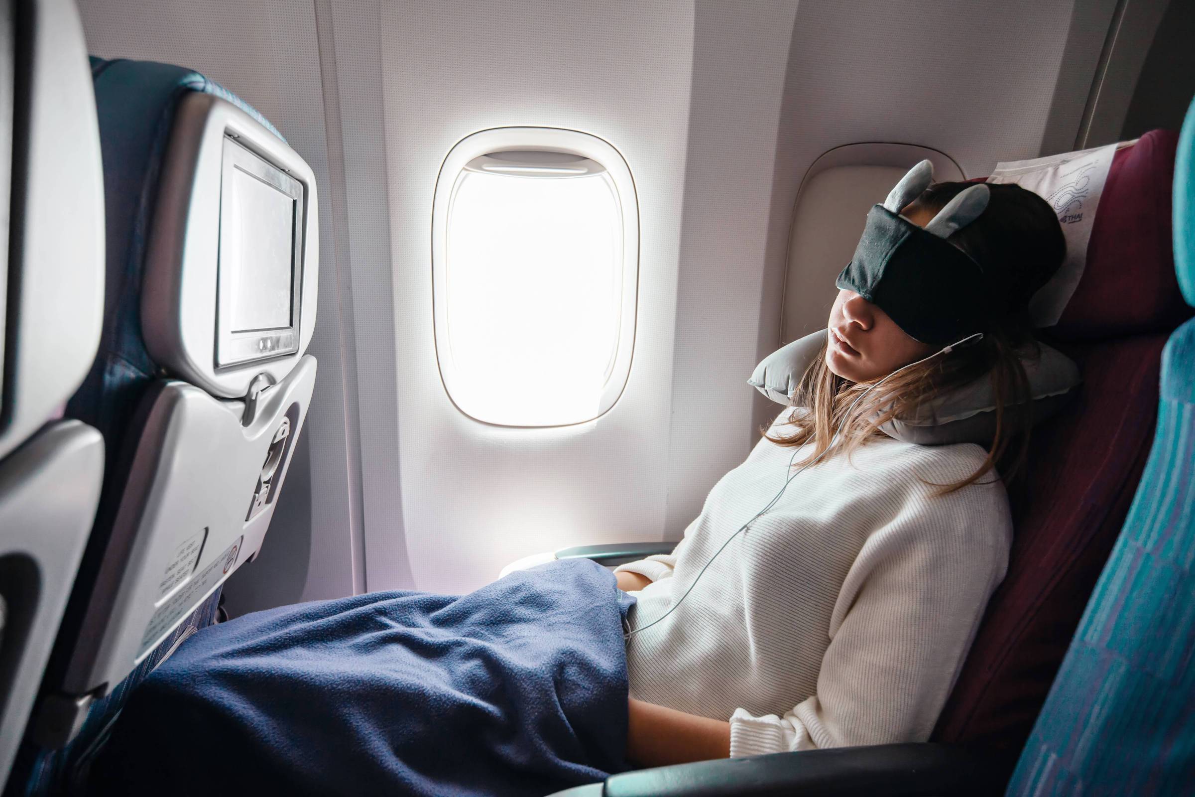Uma mulher está sentada em um assento de avião, usando uma máscara de dormir e com um travesseiro de pescoço. Ela está reclinada, com uma manta cobrindo suas pernas, e parece estar dormindo. A luz do sol entra pela janela ao lado dela, iluminando parcialmente o interior da cabine.