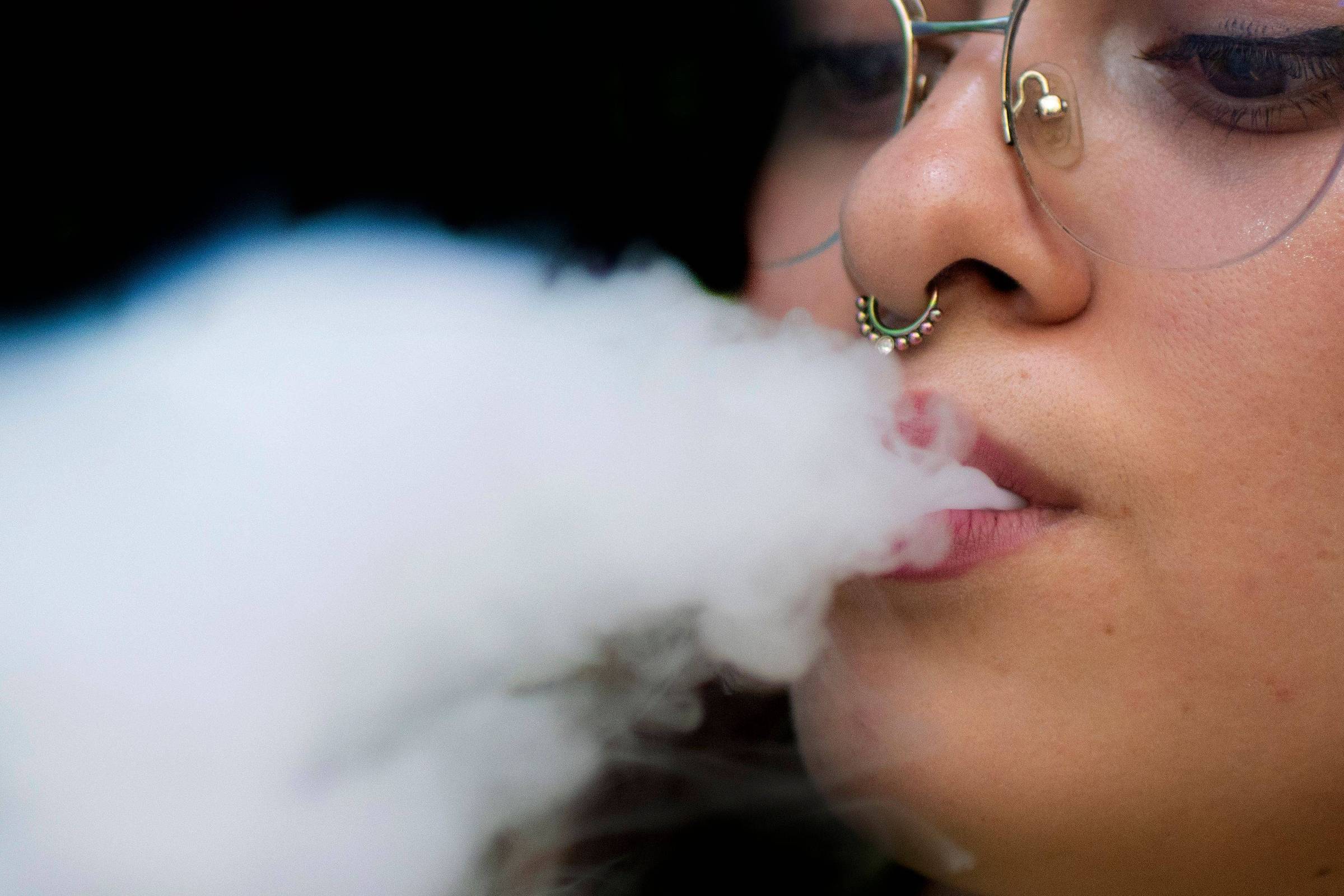 A imagem mostra uma garota jovem com óculos e um piercing no nariz exalando uma nuvem de vapor. O foco está em seu rosto, com o vapor saindo de sua boca, enquanto o fundo é desfocado.