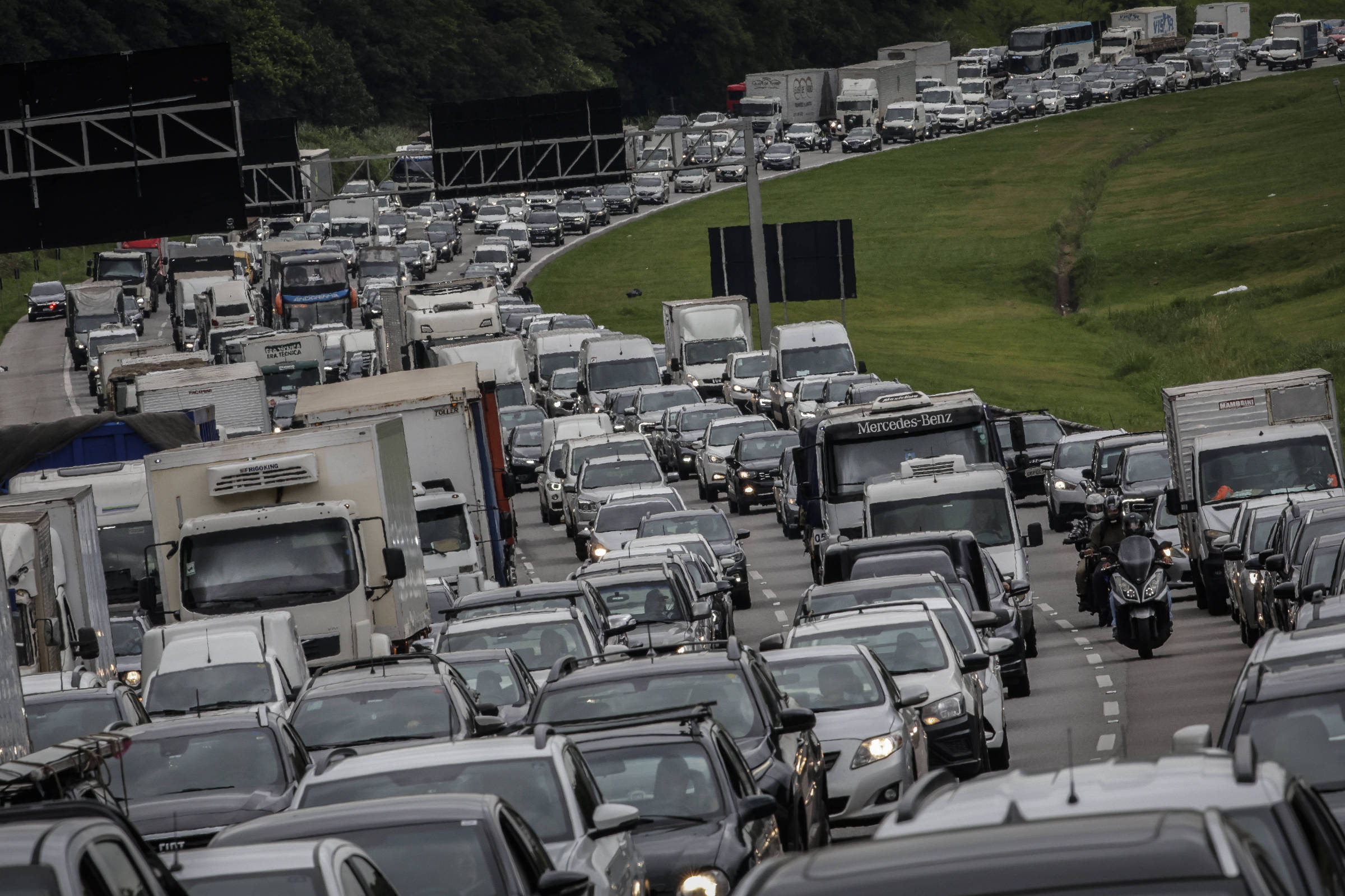 A imagem mostra um grande congestionamento em uma estrada, com muitos veículos parados, incluindo carros e caminhões. Há placas de sinalização visíveis ao fundo e a vegetação ao lado da estrada é verde. O tráfego parece intenso, com uma fila de veículos se estendendo por uma longa distância.