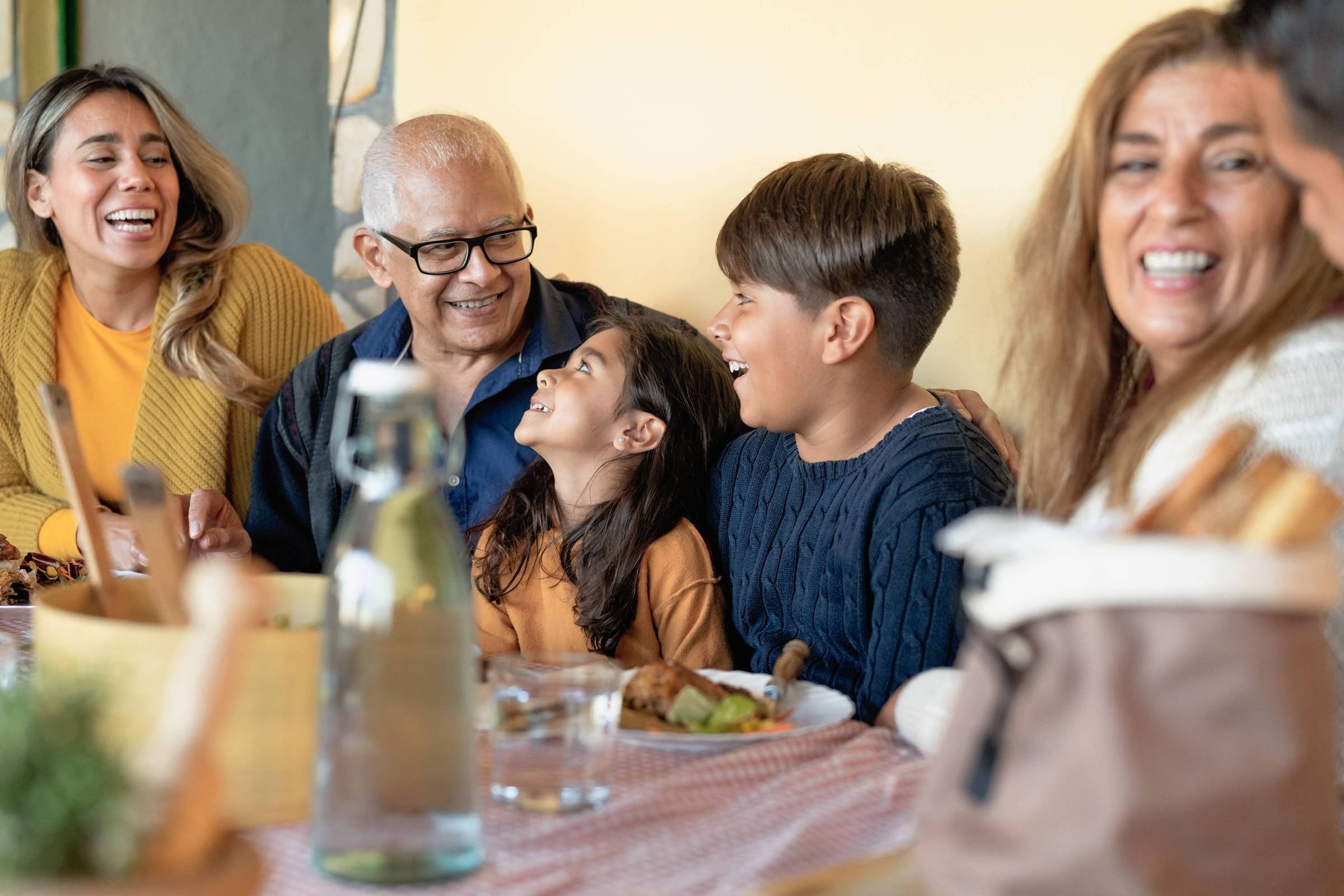 A imagem mostra uma reunião familiar em uma mesa. Há cinco pessoas sorrindo e se divertindo. Um homem idoso usa óculos e está sentado no centro, cercado por crianças e adultos. Uma menina pequena está olhando para um menino, que está rindo. As pessoas estão vestidas de maneira casual e há pratos de comida na mesa, além de uma garrafa de água. O ambiente parece acolhedor e iluminado.