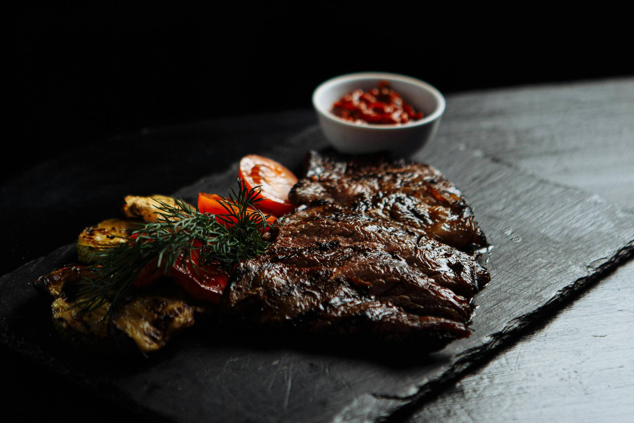 A imagem mostra um prato de carne grelhada, disposto sobre uma superfície escura. Ao lado da carne, há uma porção de legumes grelhados, incluindo pimentões e tomates. Também está presente um pequeno recipiente com molho, possivelmente barbecue, ao fundo. O ambiente é escuro, destacando os alimentos.
