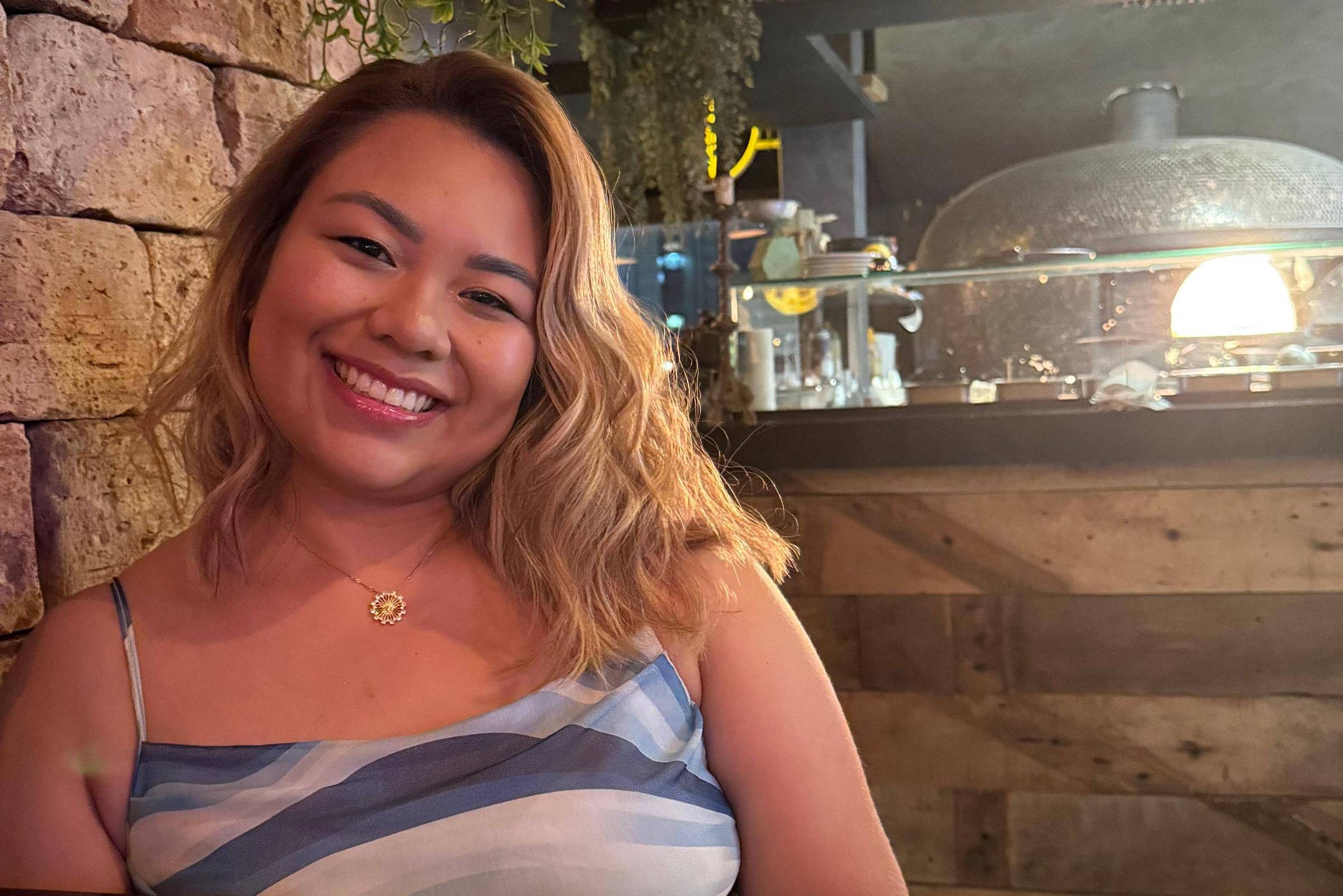 Uma jovem mulher está sentada em uma mesa de restaurante, sorrindo. Ela tem cabelo ondulado e usa uma blusa listrada em azul e branco. Ao fundo, há uma parede de pedras e uma iluminação suave, além de um forno ou equipamento de cozinha visível. Na mesa, há um cardápio.