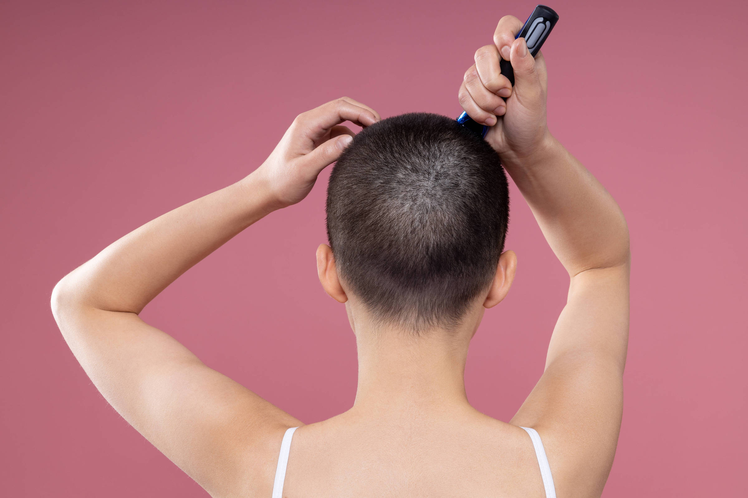 A imagem mostra a parte de trás de uma pessoa com cabelo muito curto, quase raspado, segurando uma máquina de cortar cabelo. A pessoa está de costas, com uma blusa de alças finas, e o fundo é de uma cor rosa suave. A mão esquerda está segurando a parte de trás da cabeça, enquanto a mão direita segura a máquina de cortar cabelo.