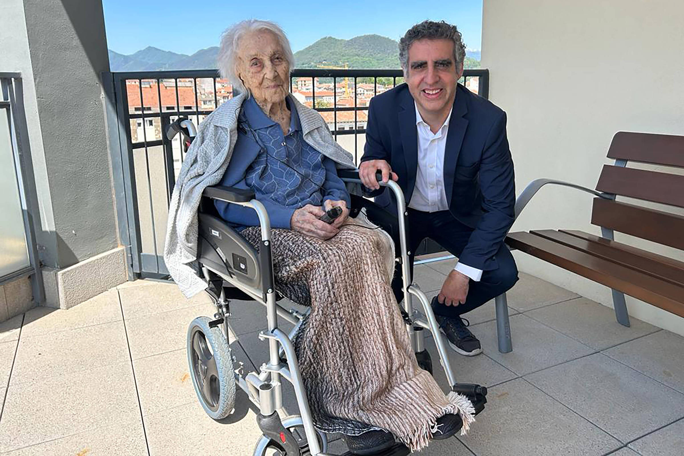 Idosa sentada em cadeira de rodas com cobertor bege, vestindo camisa jeans e casaco cinza, acompanhada por homem de terno azul escuro e camisa branca agachado ao lado dela em varanda com banco de madeira e vista de montanhas ao fundo.