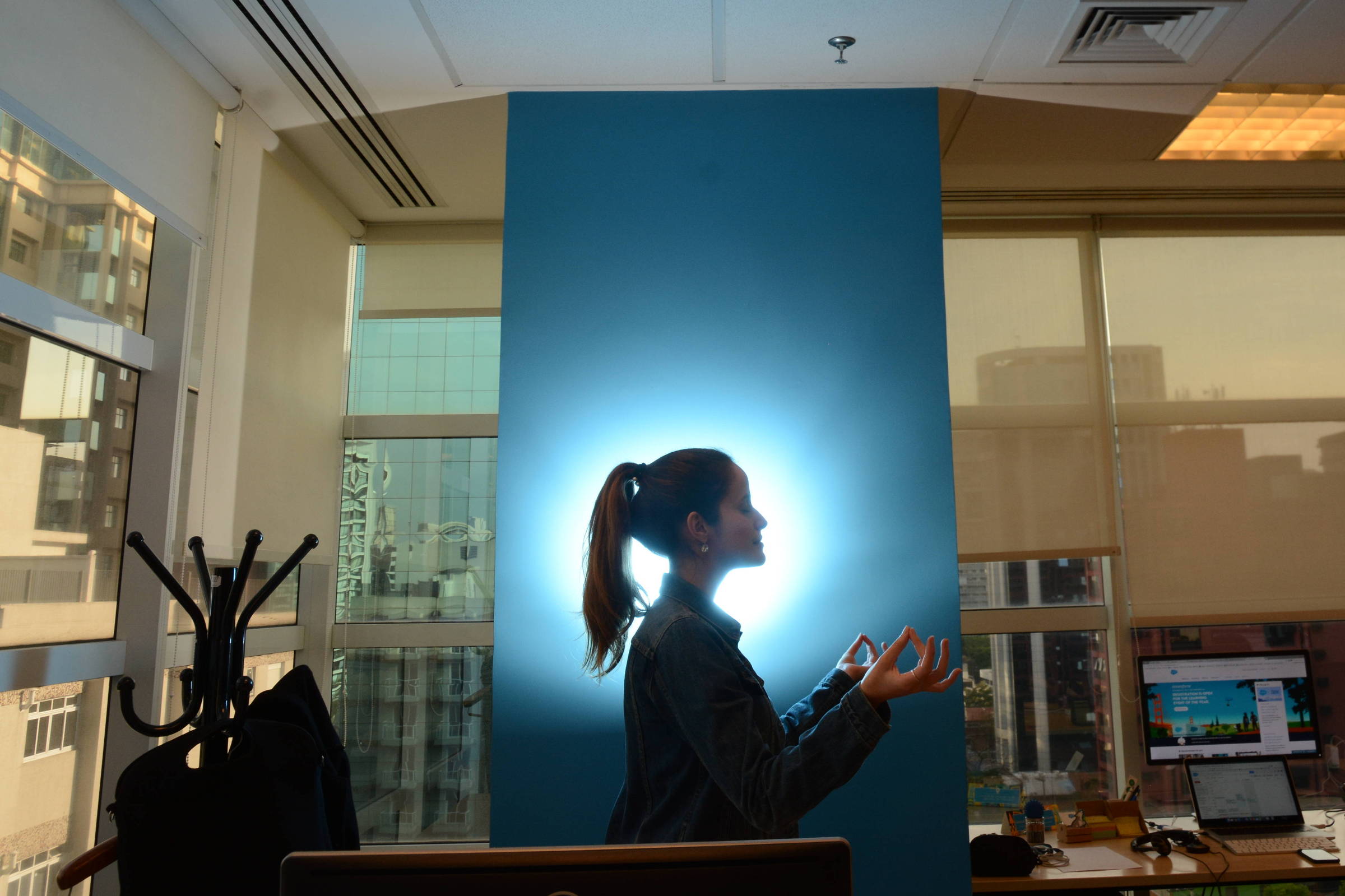 A imagem mostra a silhueta de uma mulher em um escritório, com um fundo iluminado em azul. Ela está de perfil, com os braços levantados e as palmas das mãos voltadas para cima. Ao fundo, é possível ver janelas com vista para a cidade e um ambiente de trabalho com computadores e uma cadeira