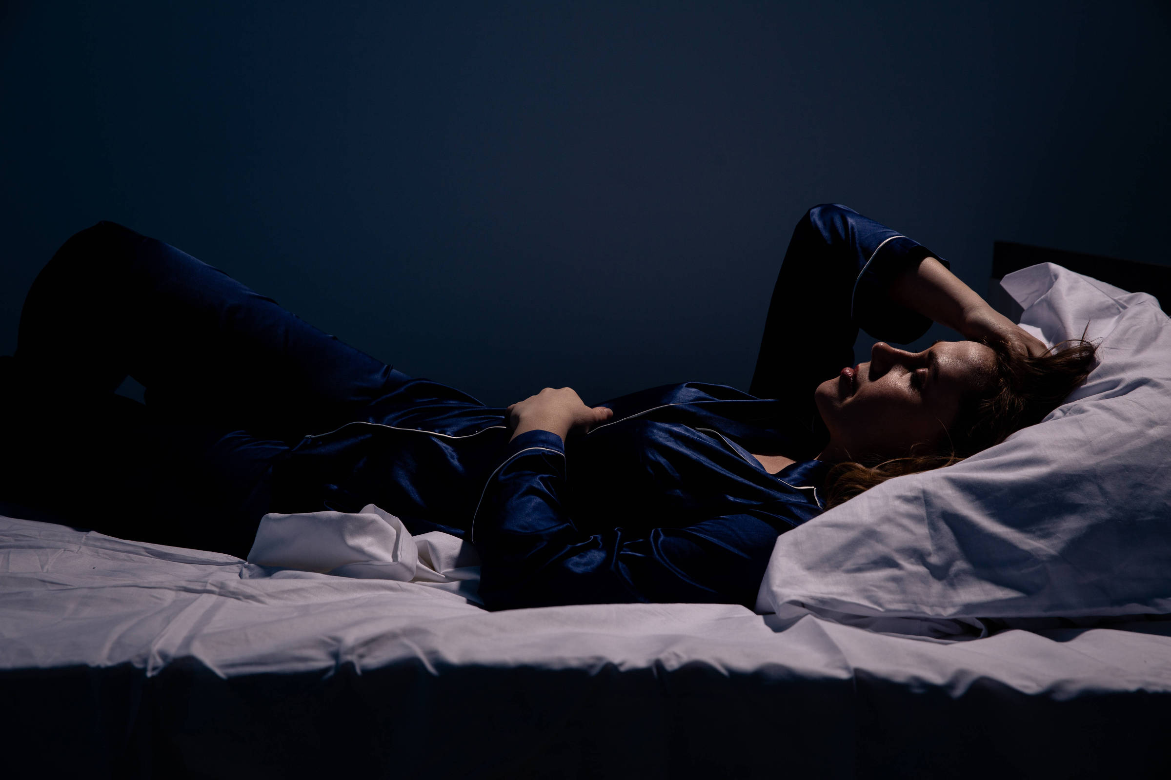 Fotografia colorida mostra uma mulher branca de pijama azul marinho deitada sobre uma cama de lençóis brancos; ela está acordada, virada de barriga para cima e com uma mão sobre a testa
