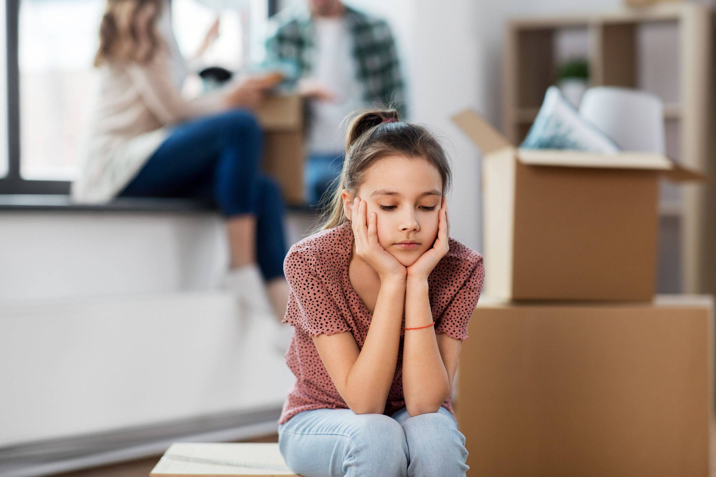 A imagem mostra uma menina com expressão triste, sentada com as mãos no rosto, em um ambiente que parece ser uma casa em processo de mudança. Ao fundo, há duas pessoas desfocadas, possivelmente adultos, interagindo perto de uma janela. Há várias caixas de papelão ao redor, indicando que a família está se mudando.
