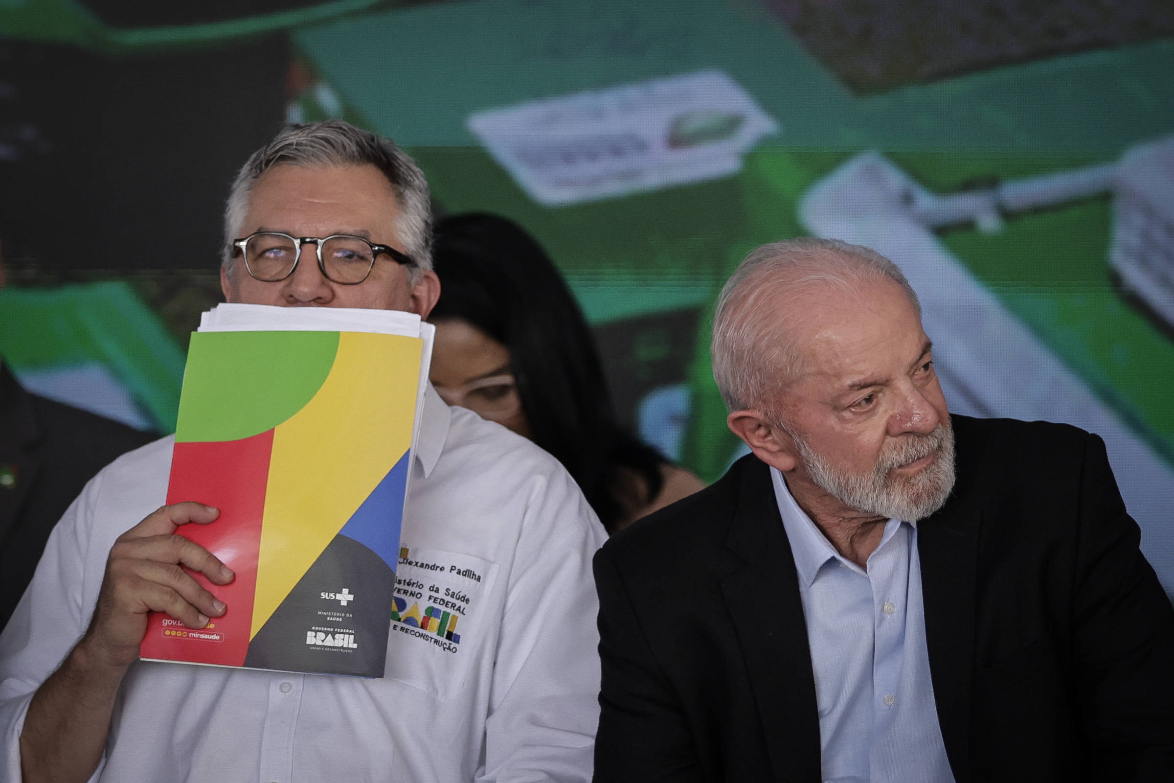 A imagem mostra duas pessoas sentadas em um evento. À esquerda, um homem com óculos e cabelo grisalho segura um documento colorido na frente do rosto. À direita, um homem mais velho com cabelo branco e barba está olhando para frente. Ao fundo, há uma tela com gráficos e informações. A atmosfera parece ser de um evento oficial ou uma conferência.