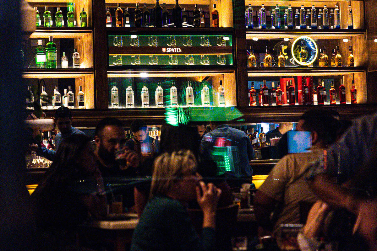 Pessoas sentadas e em pé em bar com iluminação baixa. Ao fundo, prateleiras de madeira exibem garrafas de bebidas alcoólicas organizadas por cor e tipo, com destaque para garrafas transparentes e coloridas. Ambiente interno com atmosfera de convivência noturna.