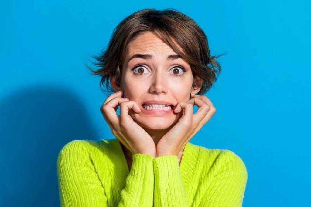 Mulher jovem de cabelos curtos castanhos usa suéter verde-limão e faz expressão facial de ansiedade, com olhos arregalados e mãos próximas à boca, sobre fundo azul.