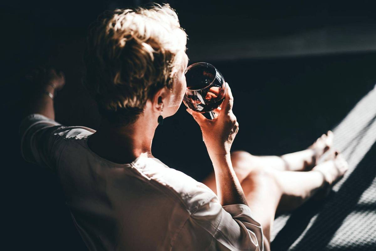 Pessoa de cabelos curtos e claros sentada, segurando uma taça de vinho tinto próxima à boca. A luz natural incide sobre o braço e as pernas, que estão estendidas, enquanto o fundo permanece escuro.