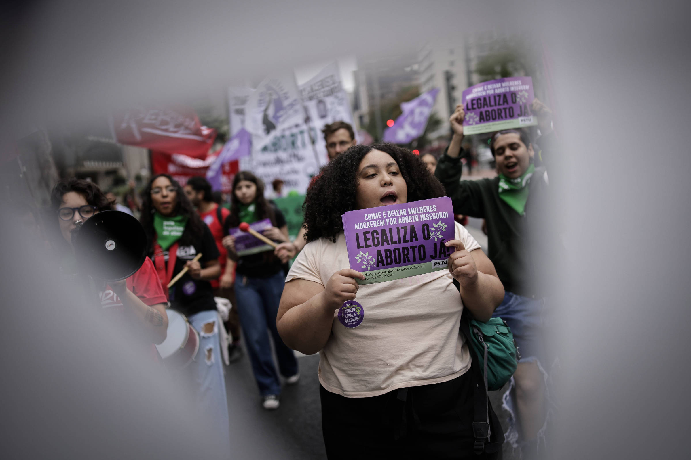 Grupo de manifestantes caminha em rua urbana segurando cartazes roxos com a frase