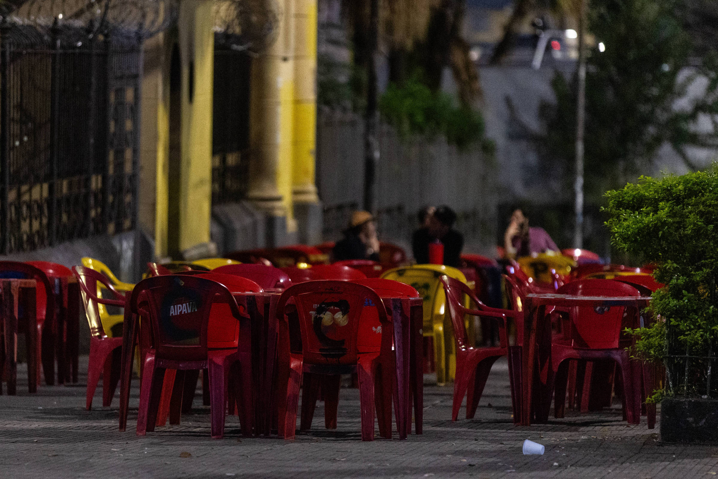 Mesas e cadeiras plásticas vermelhas e amarelas dispostas em calçada de rua à noite. Ao fundo, três pessoas sentadas conversam em ambiente externo com iluminação baixa.