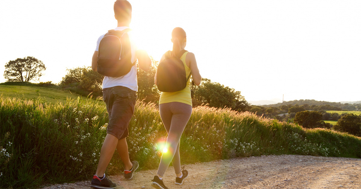 A imagem mostra duas pessoas caminhando em uma estrada de terra cercada por natureza. Elas estão de costas, andando lado a lado em direção ao pôr do sol. Ambas carregam mochilas — a pessoa à esquerda usa camiseta branca e bermuda escura, e a da direita veste regata amarela e calça legging cinza. O sol está baixo no horizonte, criando um brilho intenso e dourado que ilumina a cena. Ao redor, há campos verdes, árvores e um céu claro, transmitindo uma sensação de tranquilidade e fim de tarde.