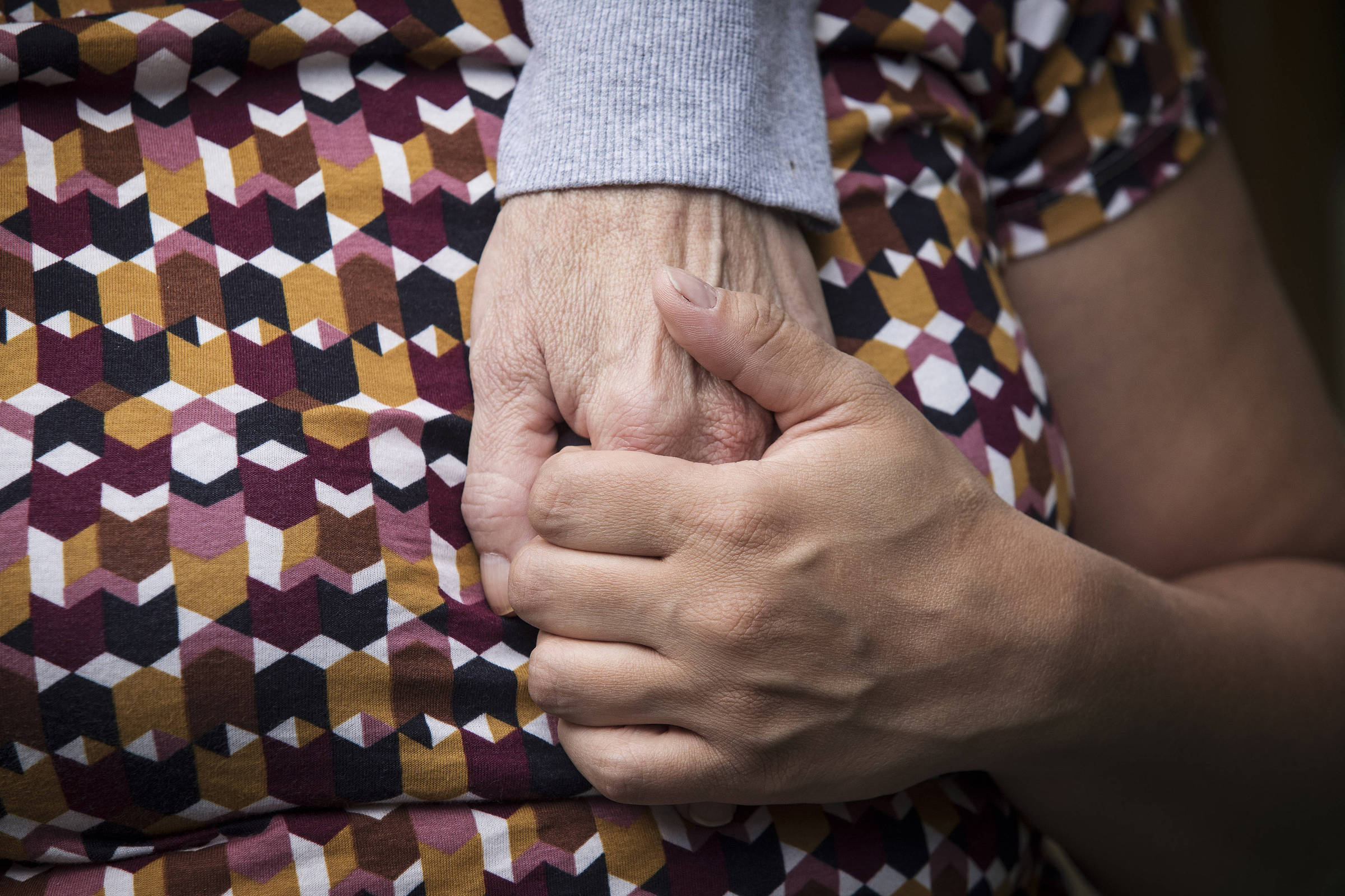 Imagem colorida foca as mãos dadas de mãe e filha; as mãos estão sobre o peito da filha