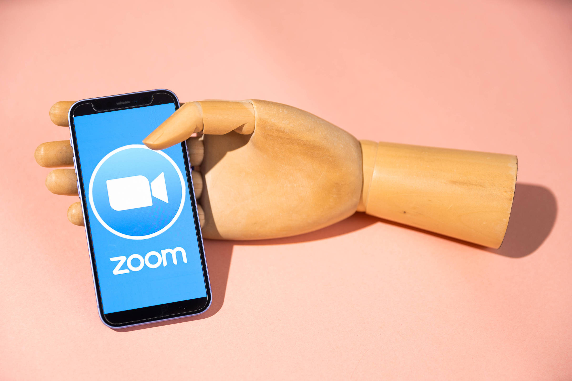 Uma mão de manequim de madeira segura um celular em que na tela está aberto o aplicativo Zoom