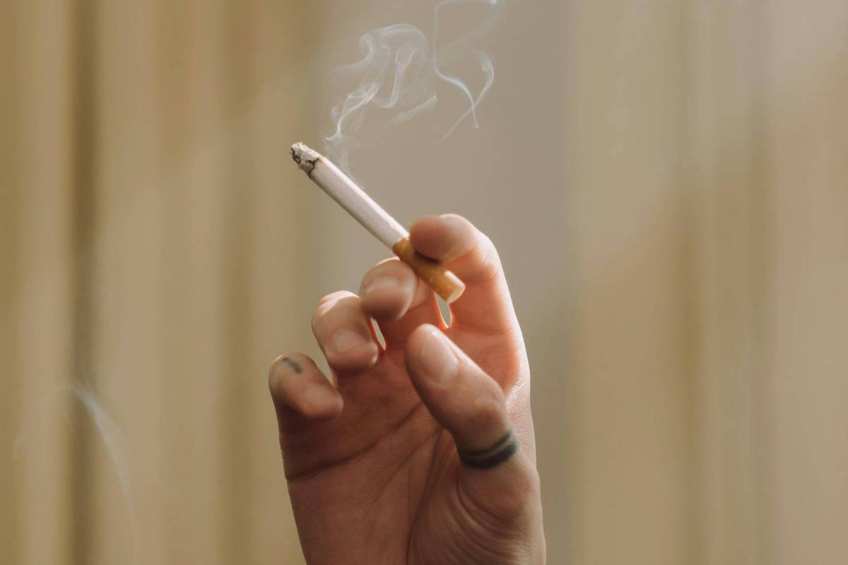 A imagem mostra uma mão segurando um cigarro aceso, com fumaça saindo dele. O fundo é desfocado, com cortinas em um tom claro, sugerindo um ambiente interno. A luz parece estar incidindo sobre a mão e o cigarro, destacando a fumaça que se eleva.