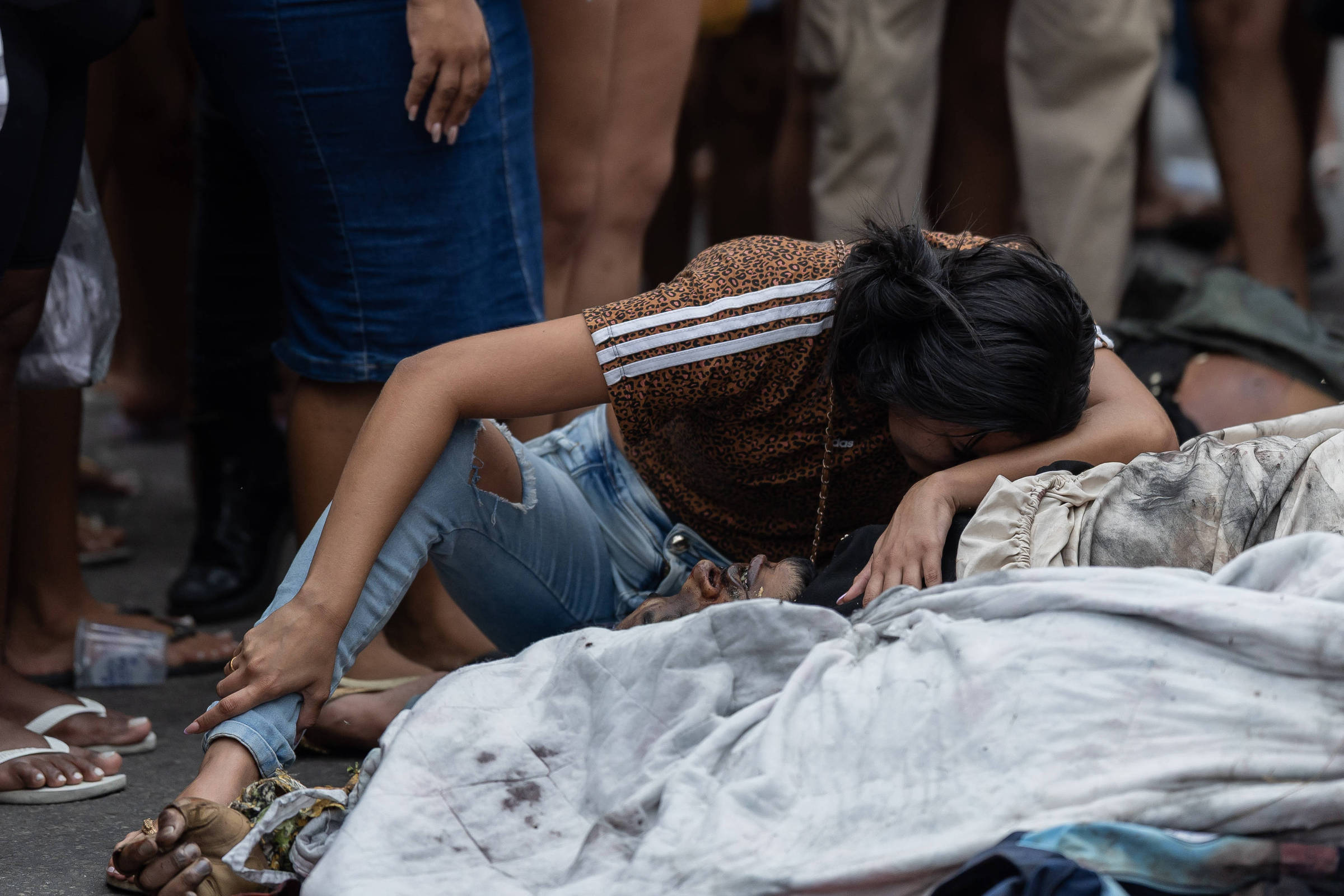 Mulher ajoelhada com camiseta marrom e calça jeans rasgada apoia a cabeça sobre corpo coberto por lençol branco manchado, cercada por pessoas em pé.