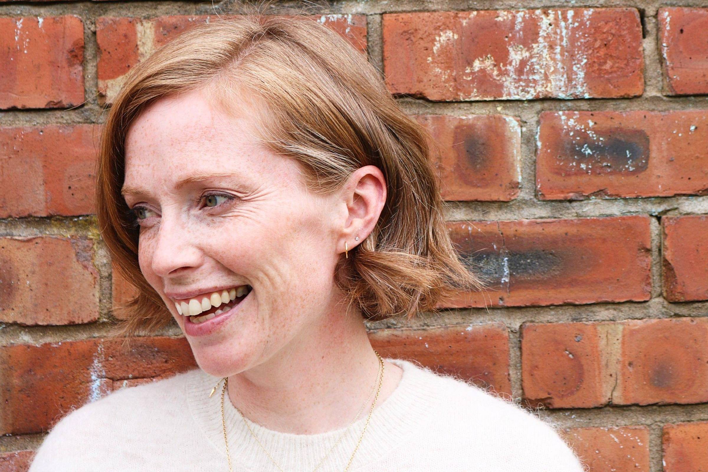 Mulher de cabelo curto loiro sorrindo e olhando para o lado, vestindo suéter branco, encostada em parede de tijolos vermelhos com marcas de desgaste.