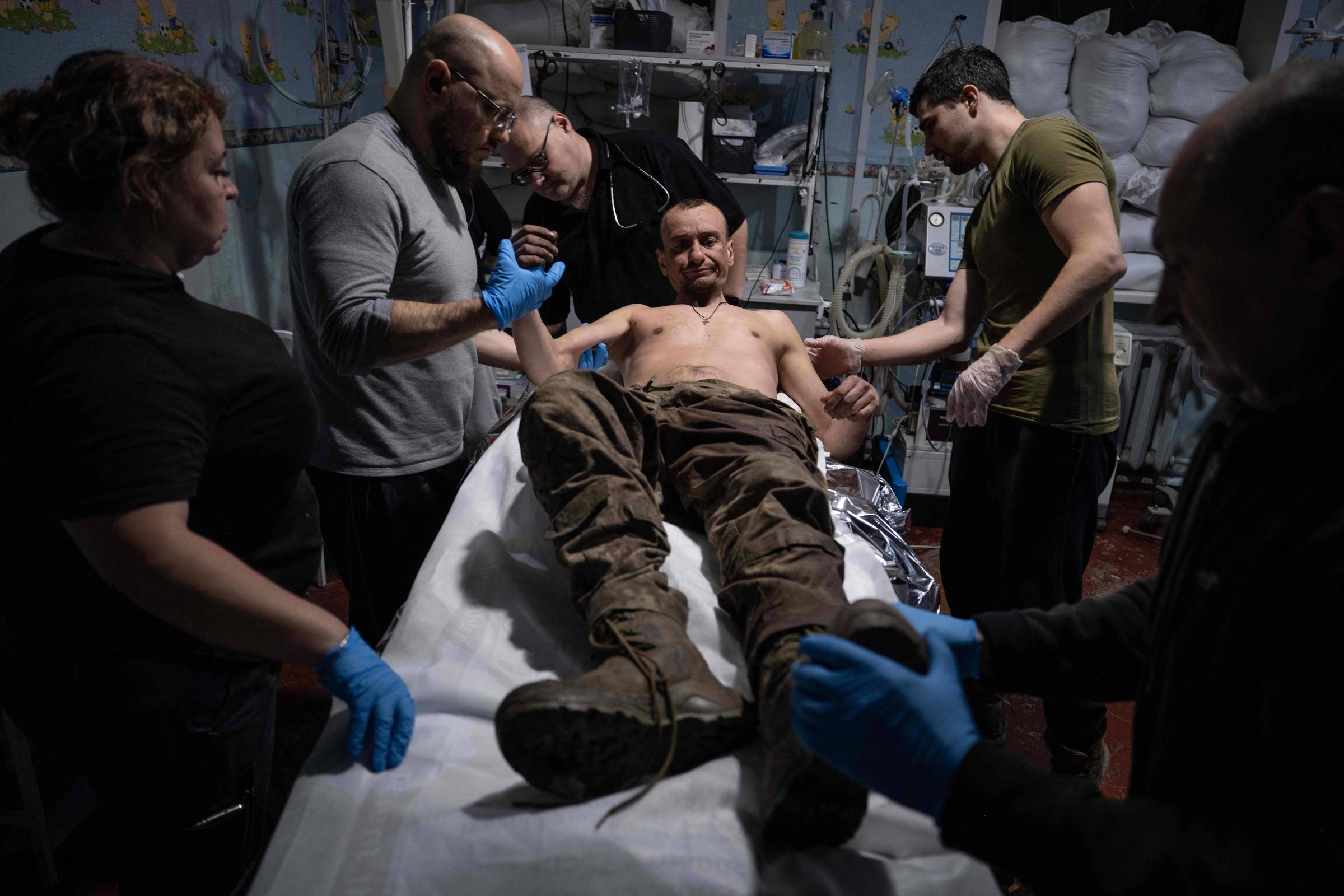 Homem sem camisa e com calça camuflada está deitado em maca enquanto quatro pessoas ao redor, usando luvas azuis, prestam atendimento médico. Equipamentos hospitalares e sacos empilhados aparecem ao fundo em sala com iluminação artificial.
