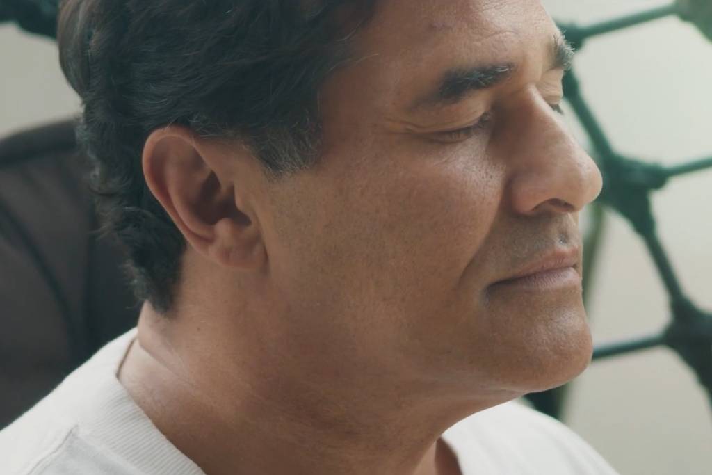 Homem de meia-idade com cabelo curto e escuro, olhos fechados, sentado em cadeira escura. Fundo desfocado com estrutura de cordas verdes.