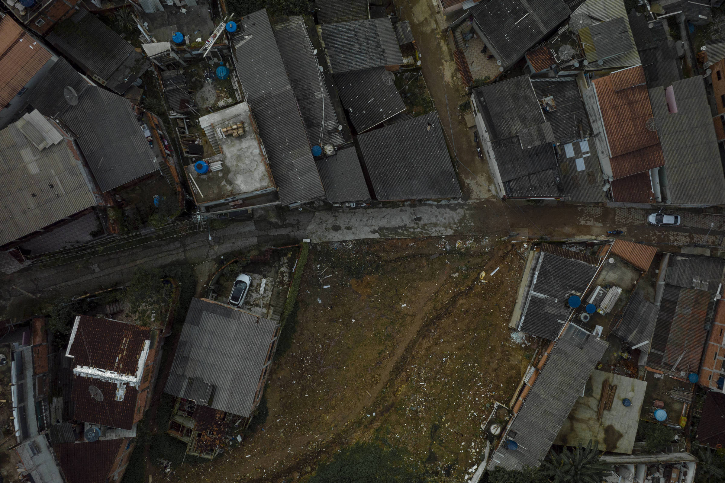 Imagem aérea mostra casas alinhadas ao redor de um terreno vazio e irregular no centro. Ruas estreitas cercam o espaço aberto, com veículos estacionados próximos às residências. Telhados variados e tanques d
