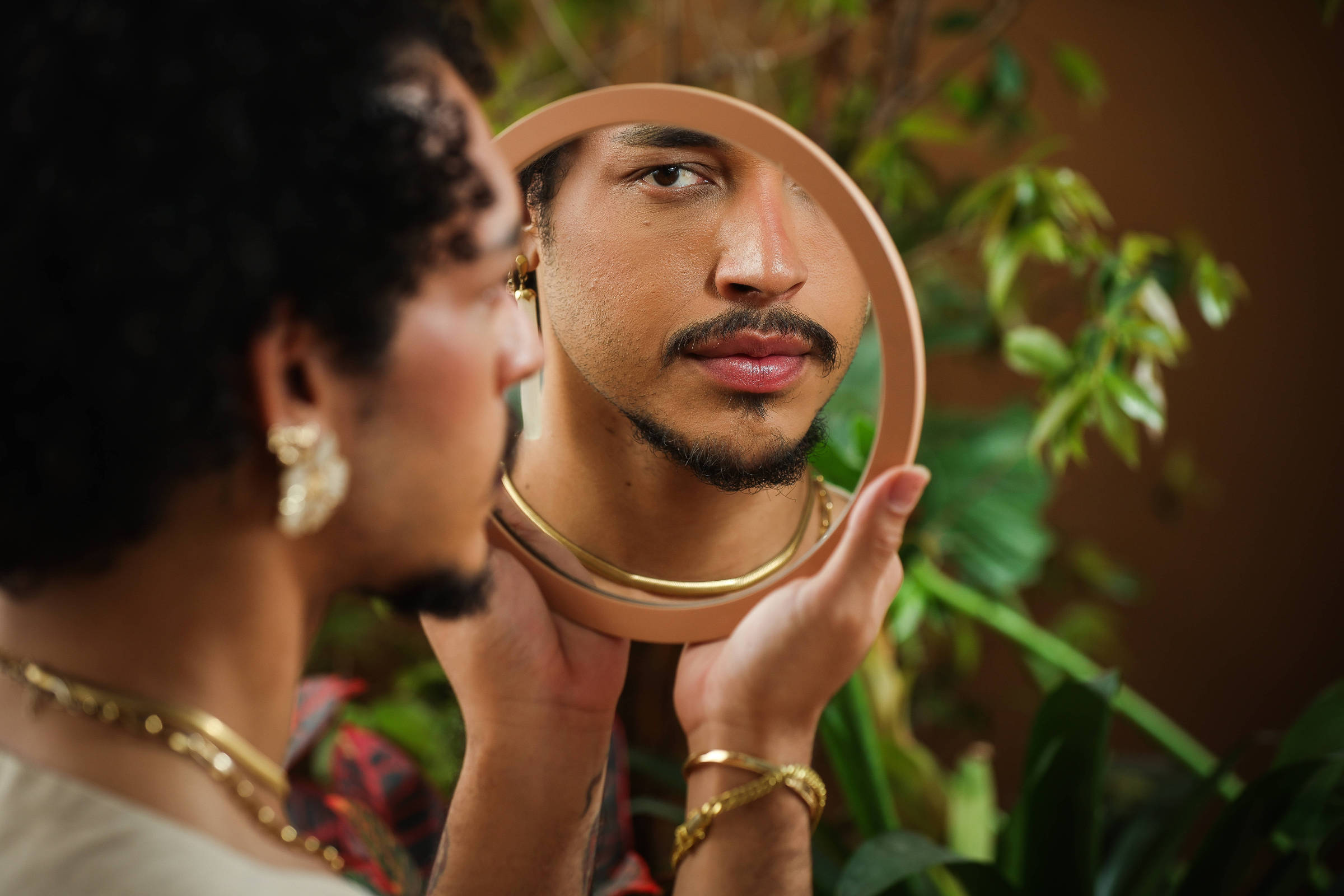 Homem com cabelo cacheado e acessórios dourados segura espelho redondo refletindo seu rosto com expressão neutra. Fundo com plantas verdes desfocadas.