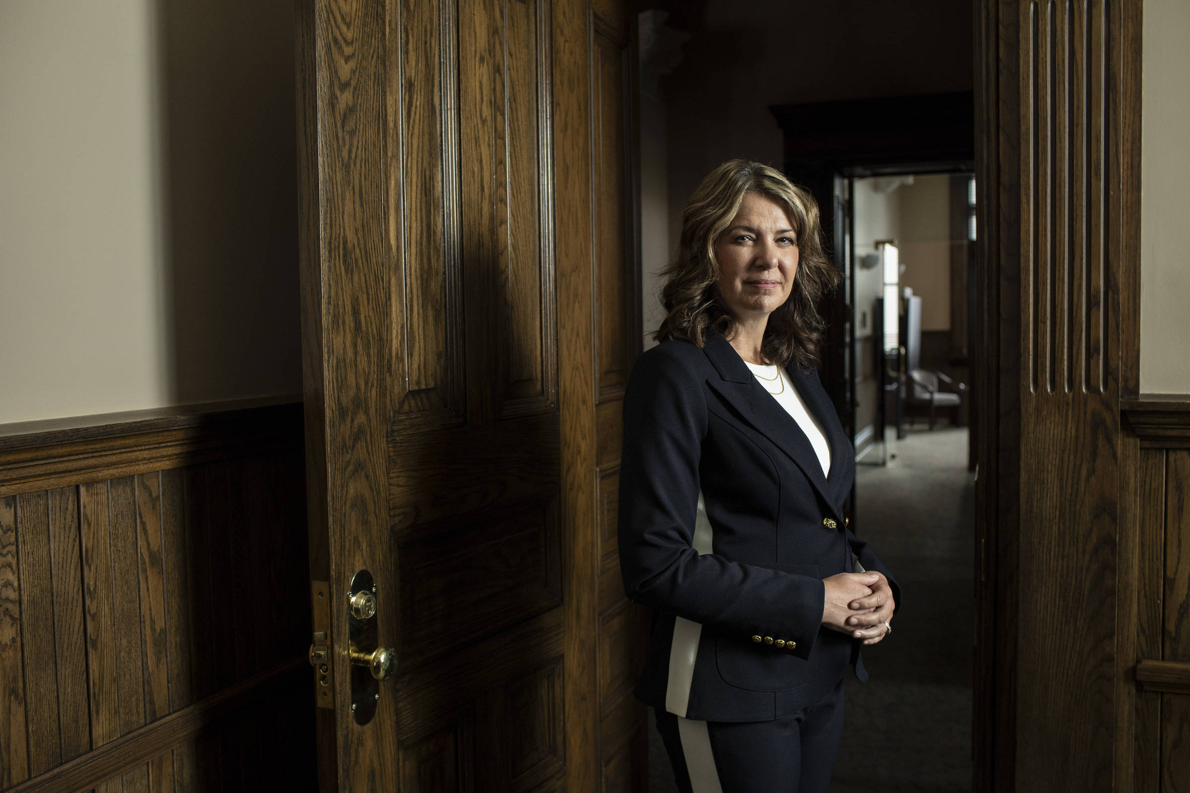 Mulher de cabelos ondulados e loiros veste terno azul escuro com detalhes claros, está encostada em porta de madeira aberta em corredor interno com paredes e portas de madeira.