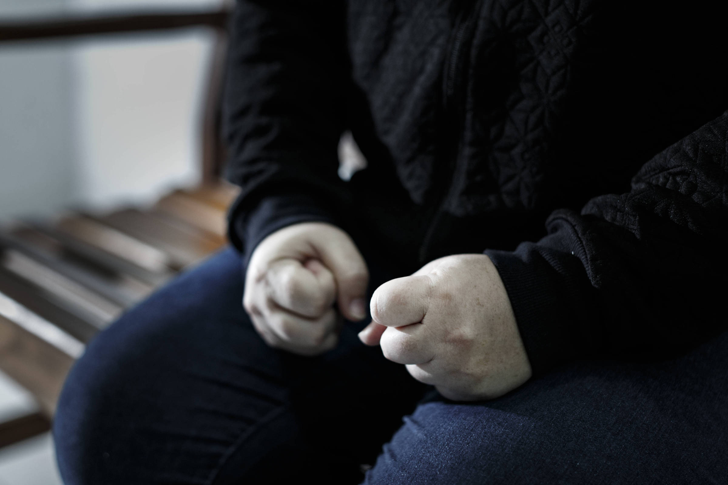 Fotografia colorida mostra uma pessoa sentada em um banco de madeira; a imagem destaca apenas parte dos braços, que estão com os punhos cerrados sobre as pernas; ela veste uma blusa preta de mangas compridas e uma calça jeans