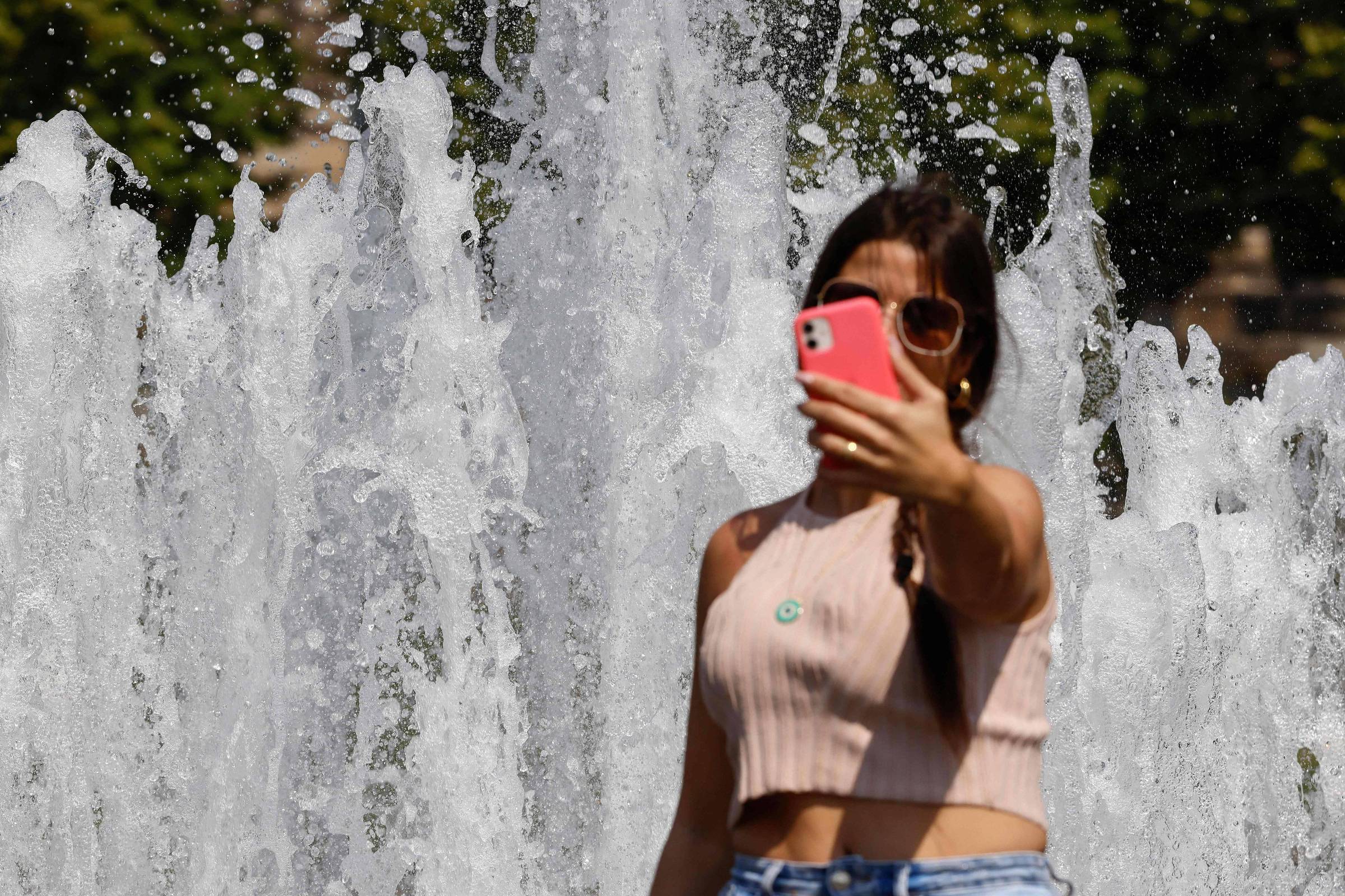 Uma mulher de cabelos lisos e pretos tira uma selfie com o celular em frete a um chafariz