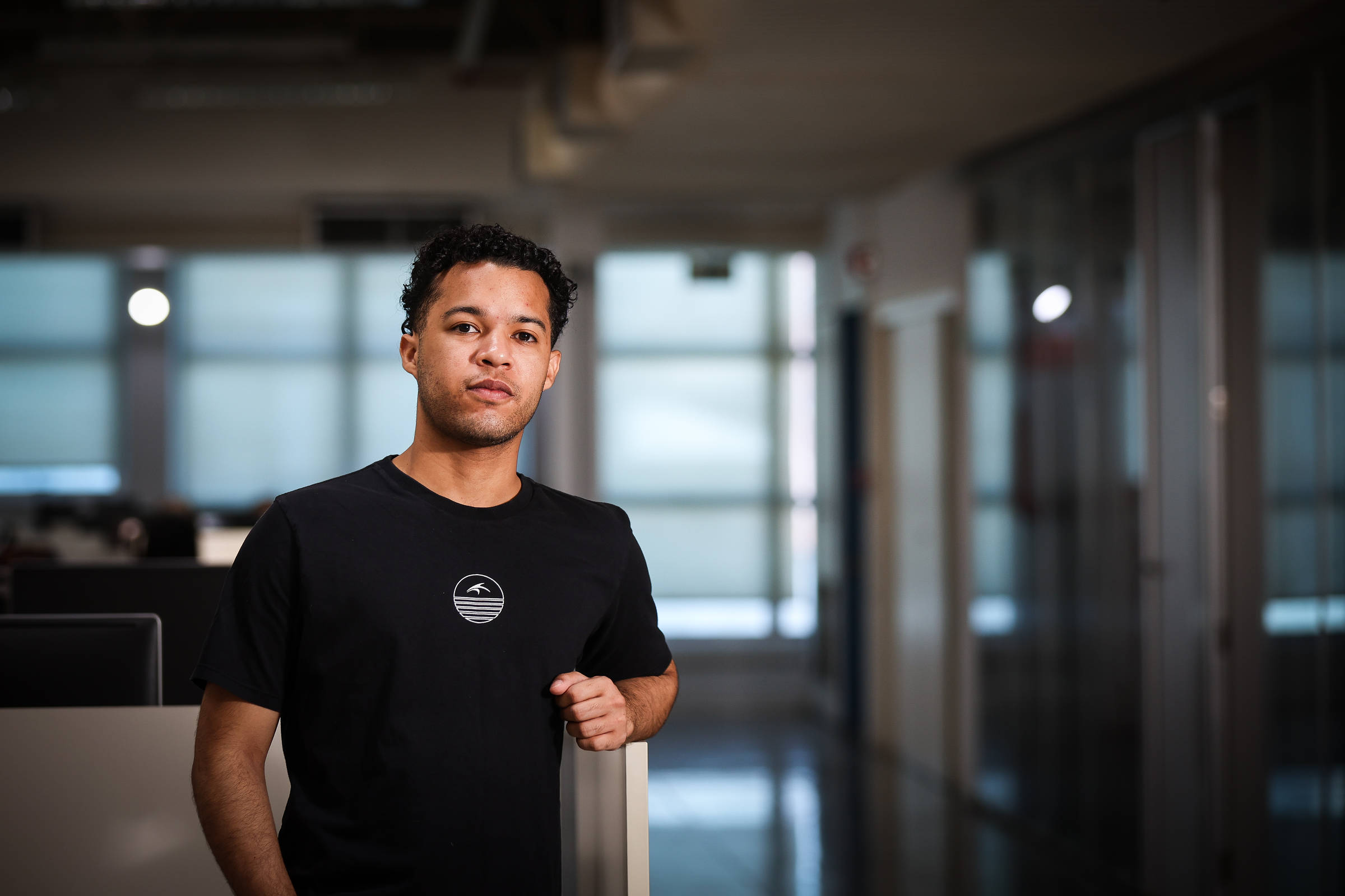 Homem jovem de camiseta preta está encostado em divisória de escritório com iluminação natural ao fundo. Espaço amplo com paredes de vidro e corredores visíveis.