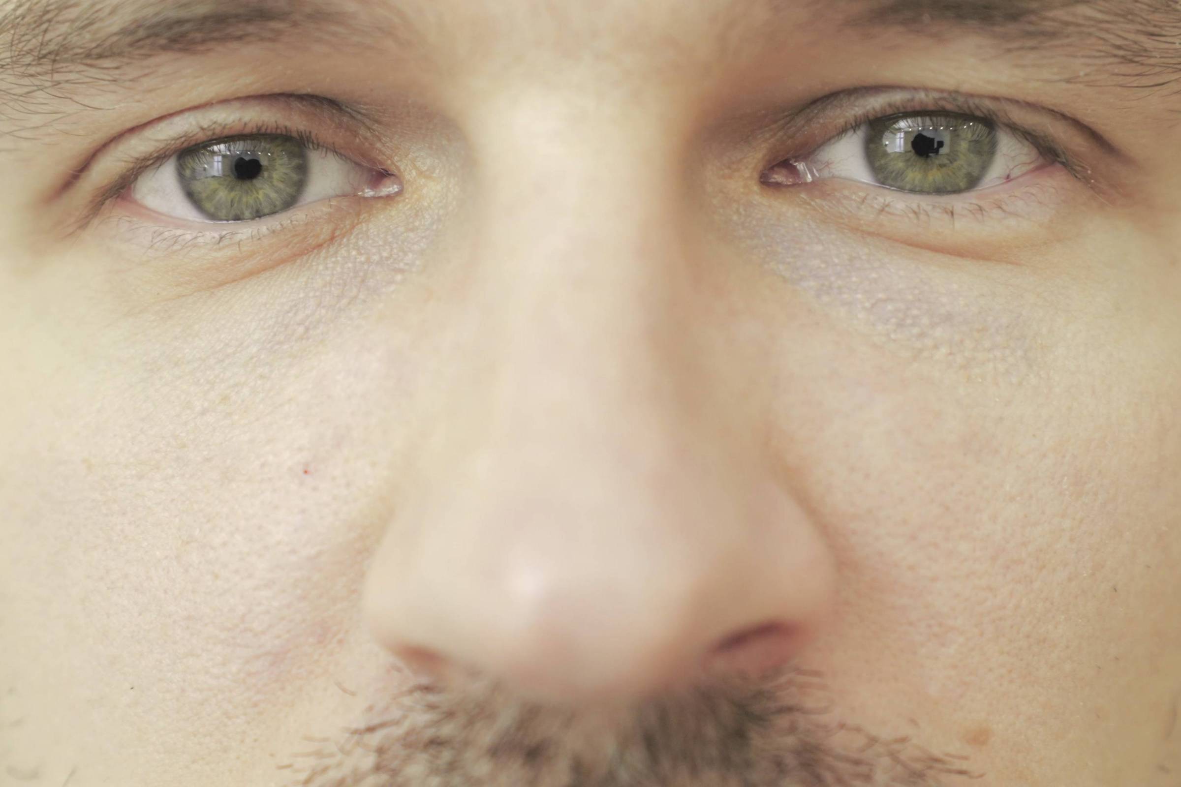 Close no rosto de um homem com olhos verdes, pele clara e barba rala. A imagem foca principalmente nos olhos e nariz.