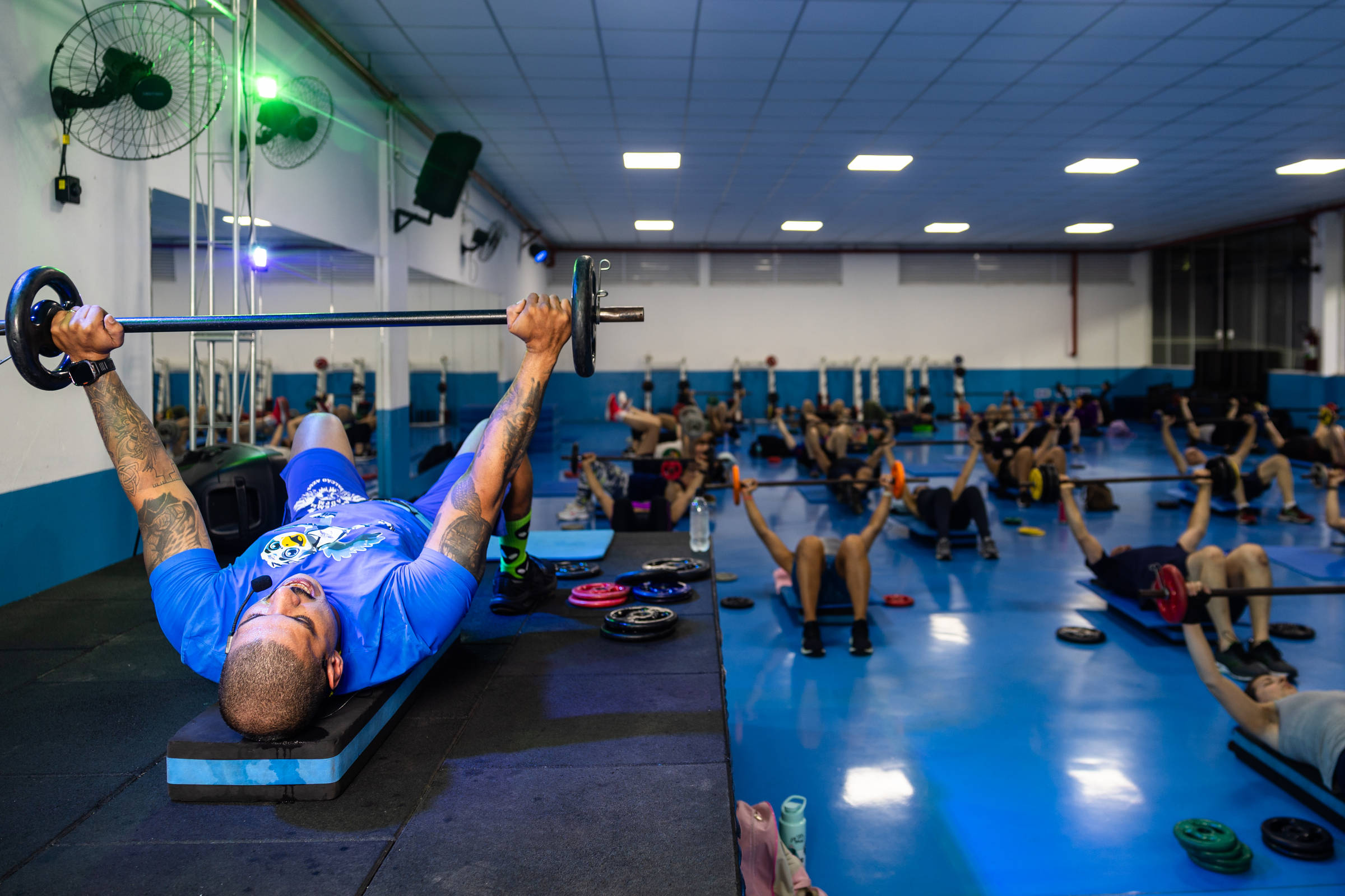 Homem de camiseta azul realiza supino com barra na frente de grupo em academia. Várias pessoas deitadas no chão fazem exercícios com halteres em ambiente iluminado e espaçoso.