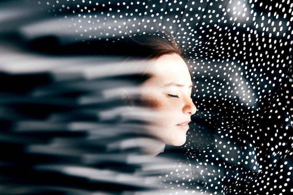 Perfil de mulher de olhos fechados com expressão serena, cercada por pontos brancos dispersos em fundo escuro, com efeito de movimento desfocado na lateral esquerda.