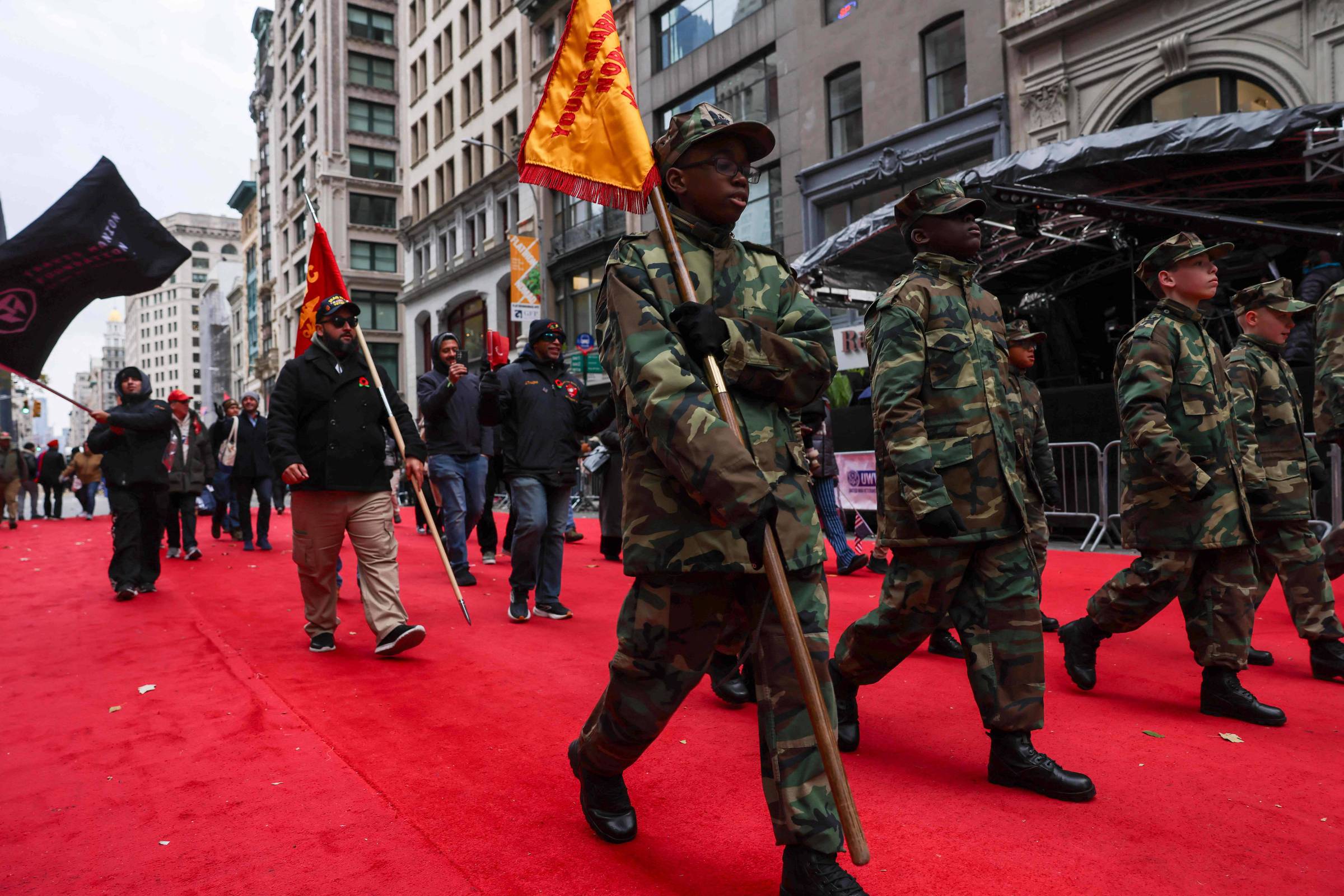 Quatro pessoas vestindo uniformes militares camuflados marcham em fila sobre um tapete vermelho em rua de área urbana. Atrás deles, outras pessoas caminham carregando bandeiras vermelhas e pretas. Edifícios altos e fachadas comerciais cercam a cena.