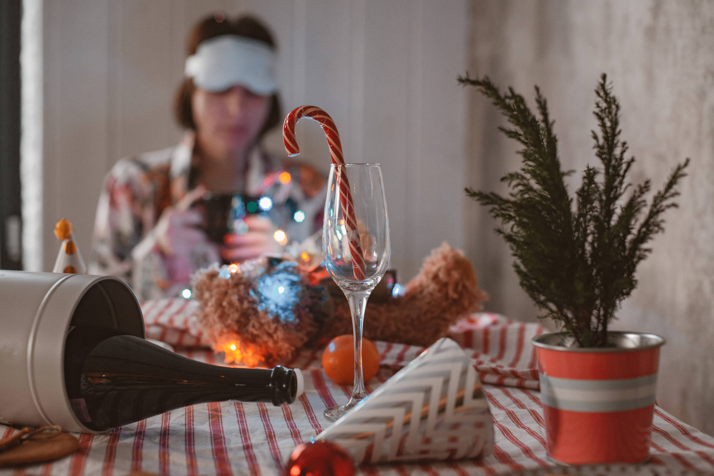 Mesa com toalha listrada exibe taça com bengala doce, garrafa de vinho caída, chapéu de festa e vaso com planta. Ao fundo, pessoa desfocada usa máscara de dormir e segura câmera.
