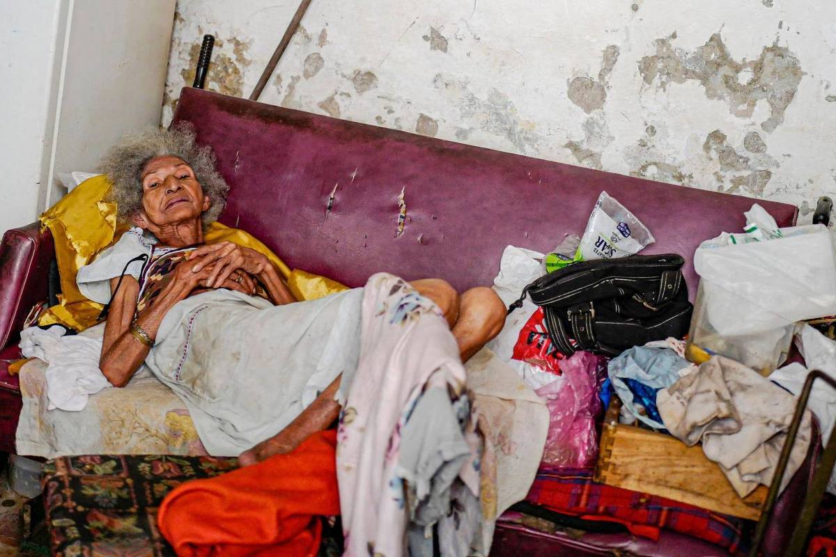 Mulher idosa de cabelos grisalhos está deitada em um sofá vermelho desgastado, com cobertores e roupas ao redor. A parede ao fundo apresenta manchas e descascados, e há objetos pessoais e sacolas próximas a ela.