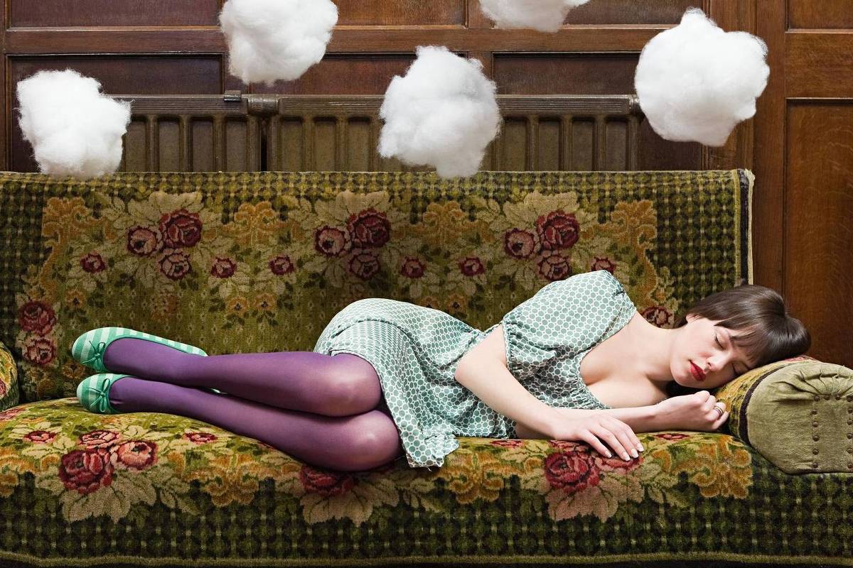 Mulher jovem deitada de lado em sofá verde com estampa floral, vestindo vestido verde claro e meia-calça roxa. Acima dela, cinco nuvens de algodão suspensas no ar, em ambiente interno com móveis de madeira ao fundo.