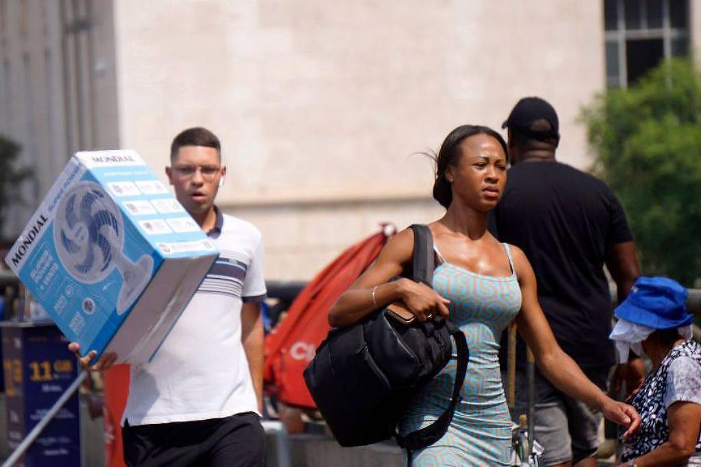 Mulher com vestido azul claro e bolsa preta caminha em calçada urbana. Homem atrás dela carrega caixa grande de ventilador. Pessoas sentadas e edifícios ao fundo.