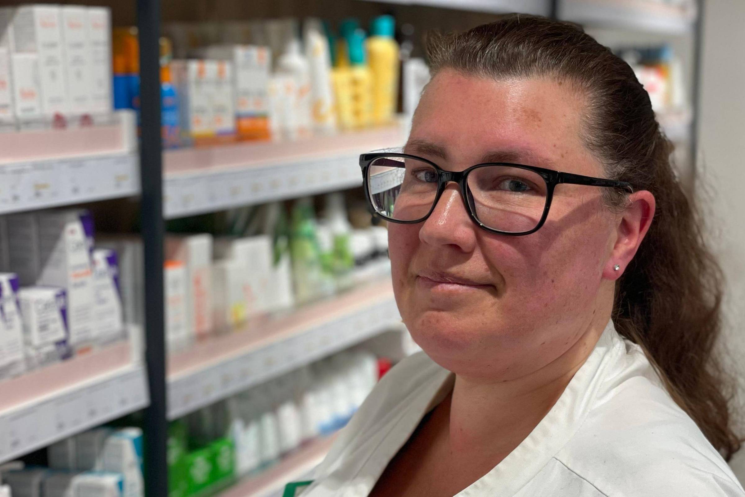 Mulher com jaleco branco e óculos está em uma farmácia, com prateleiras cheias de medicamentos e produtos ao fundo.