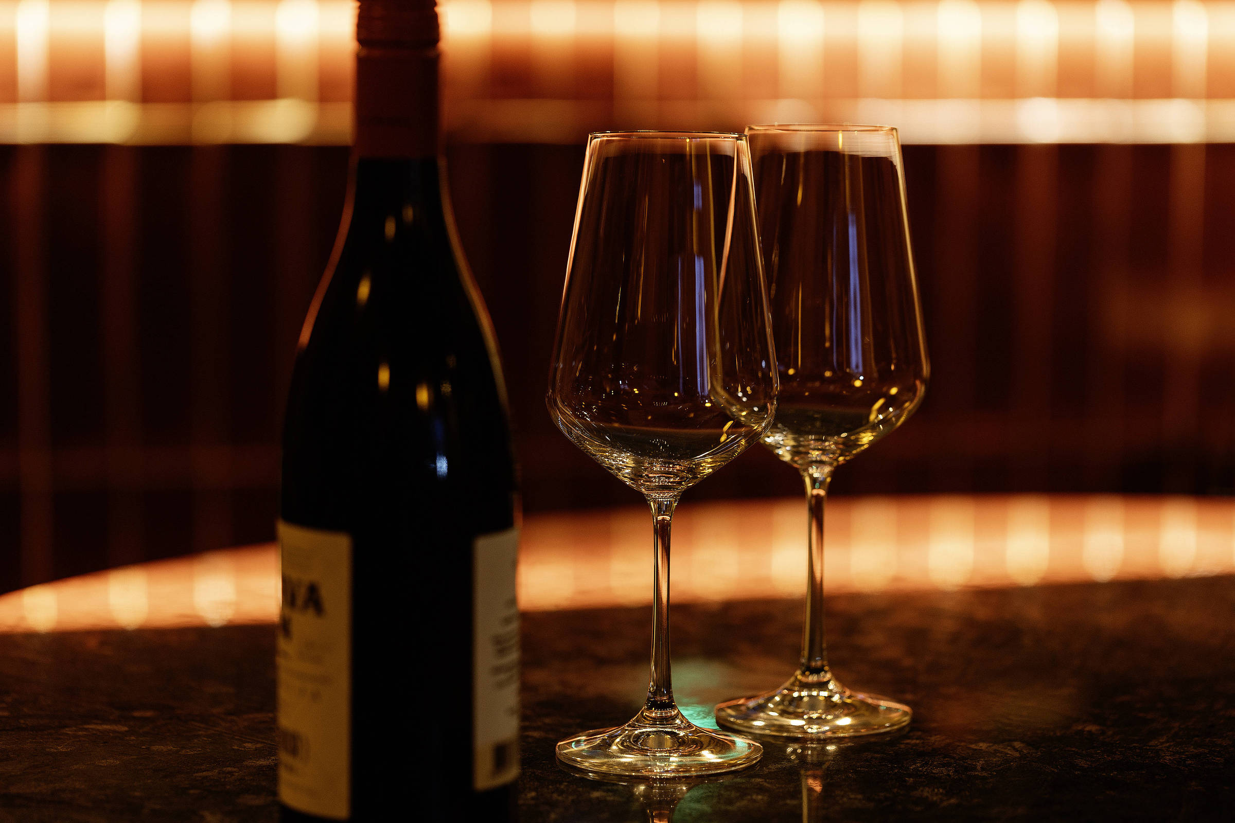 A imagem mostra uma garrafa de vinho tinto ao lado de duas taças de vinho vazias. O fundo é iluminado com luzes quentes, criando um ambiente aconchegante. A superfície onde os objetos estão apoiados é escura e reflexiva.