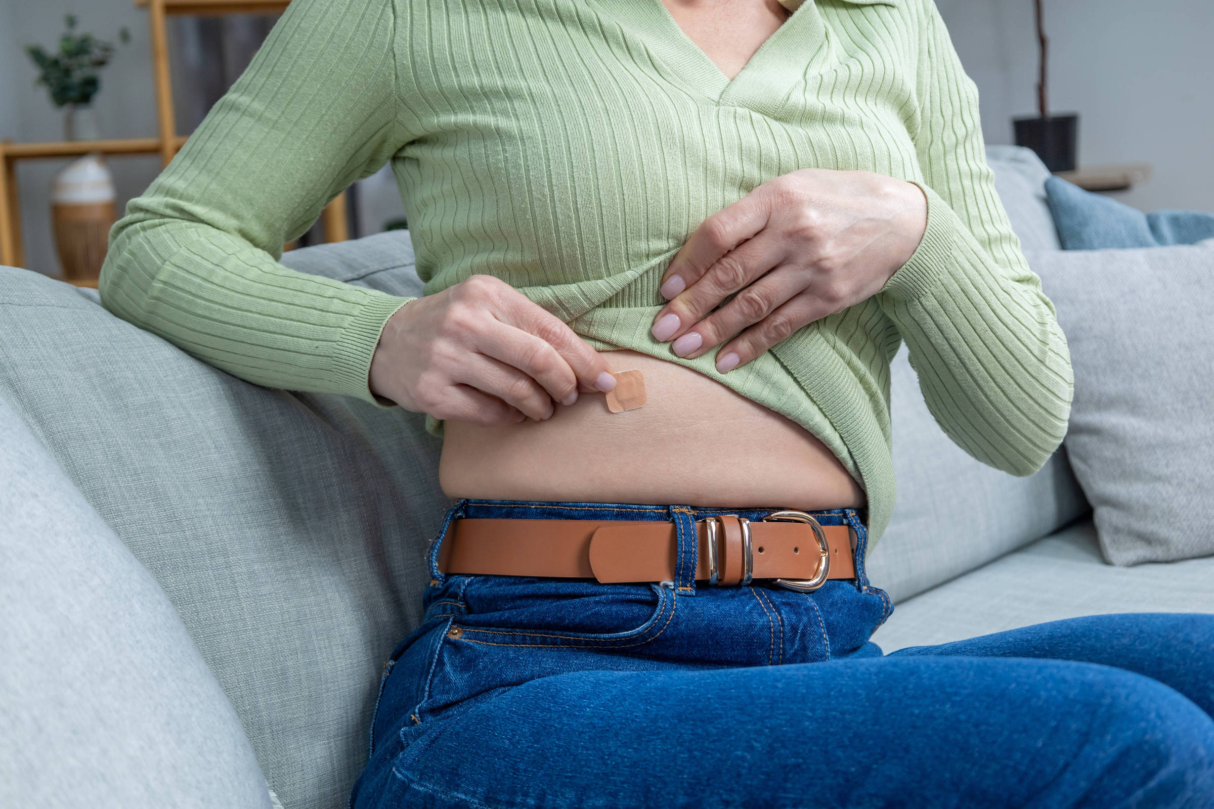 Pessoa sentada em sofá cinza levanta a blusa verde para aplicar curativo adesivo no abdômen. Veste calça jeans azul com cinto marrom.