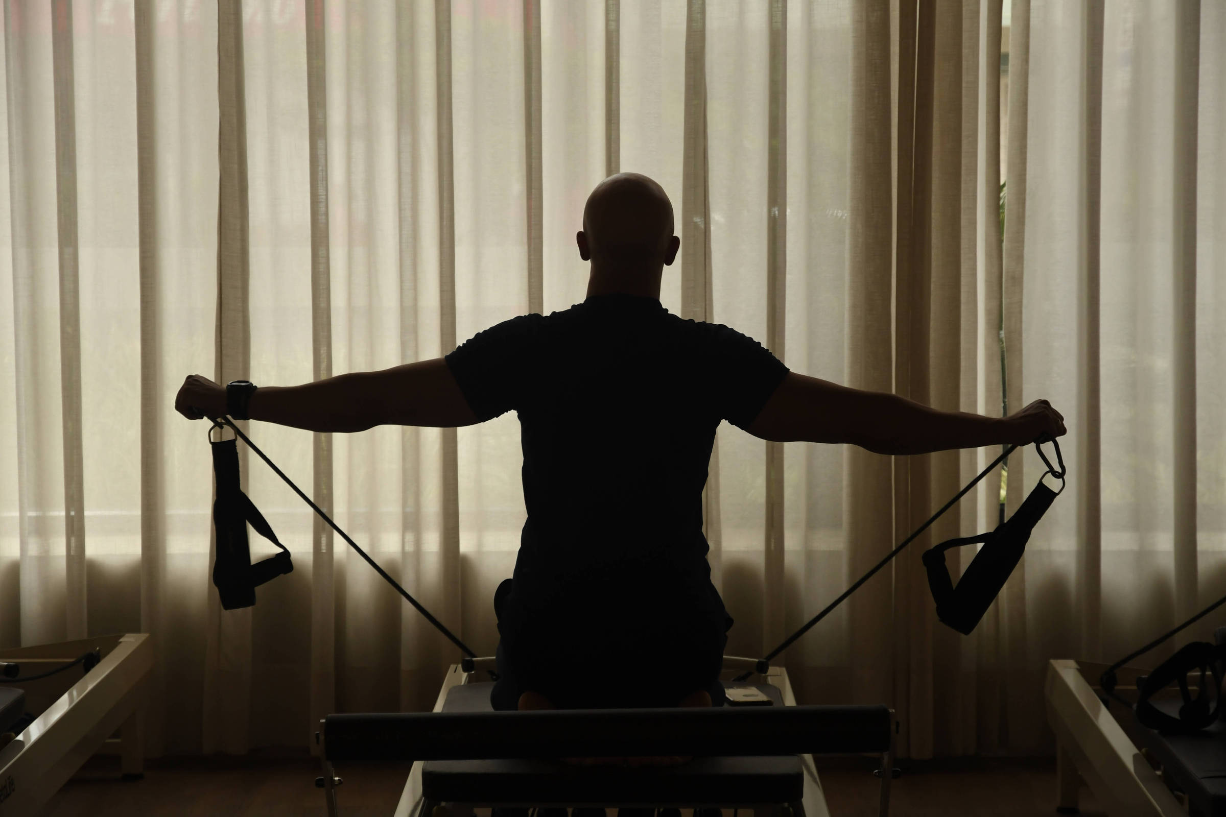 Pessoa vista de costas, sentada em aparelho de pilates, estende os braços segurando alças de resistência. Cortina translúcida ao fundo deixa a luz natural entrar no ambiente.