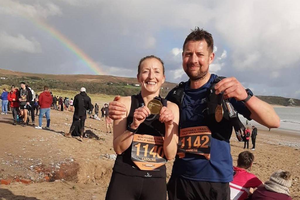 Homem e mulher sorridentes seguram medalhas e usam números de corrida na praia. Pessoas e um arco-íris aparecem ao fundo sob céu nublado.